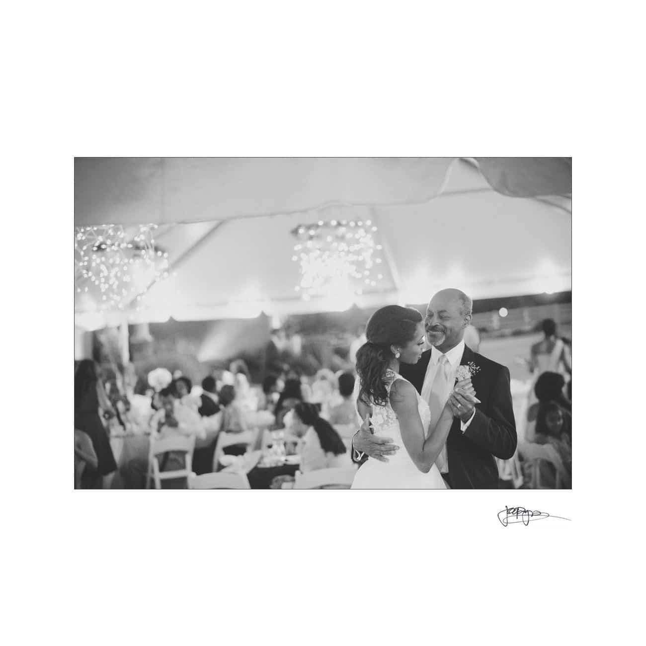 A bride and groom share a dance under a tent with string lights, surrounded by seated guests at tables. The scene is captured in black and white, highlighting the couples joyful expressions.
