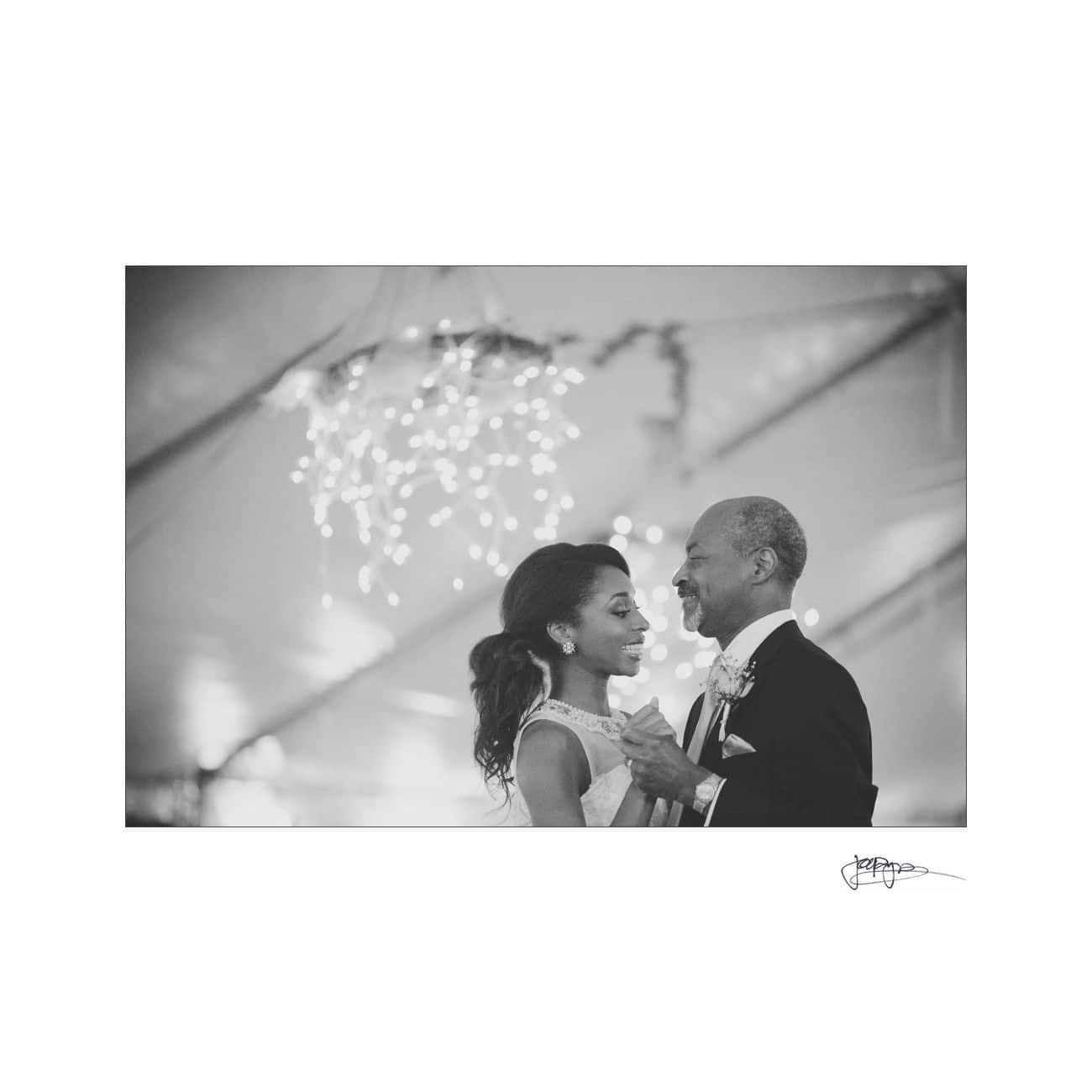 A black and white photo of a bride and an older man dancing together under a canopy with string lights. Both are smiling warmly, creating a joyful, intimate atmosphere.