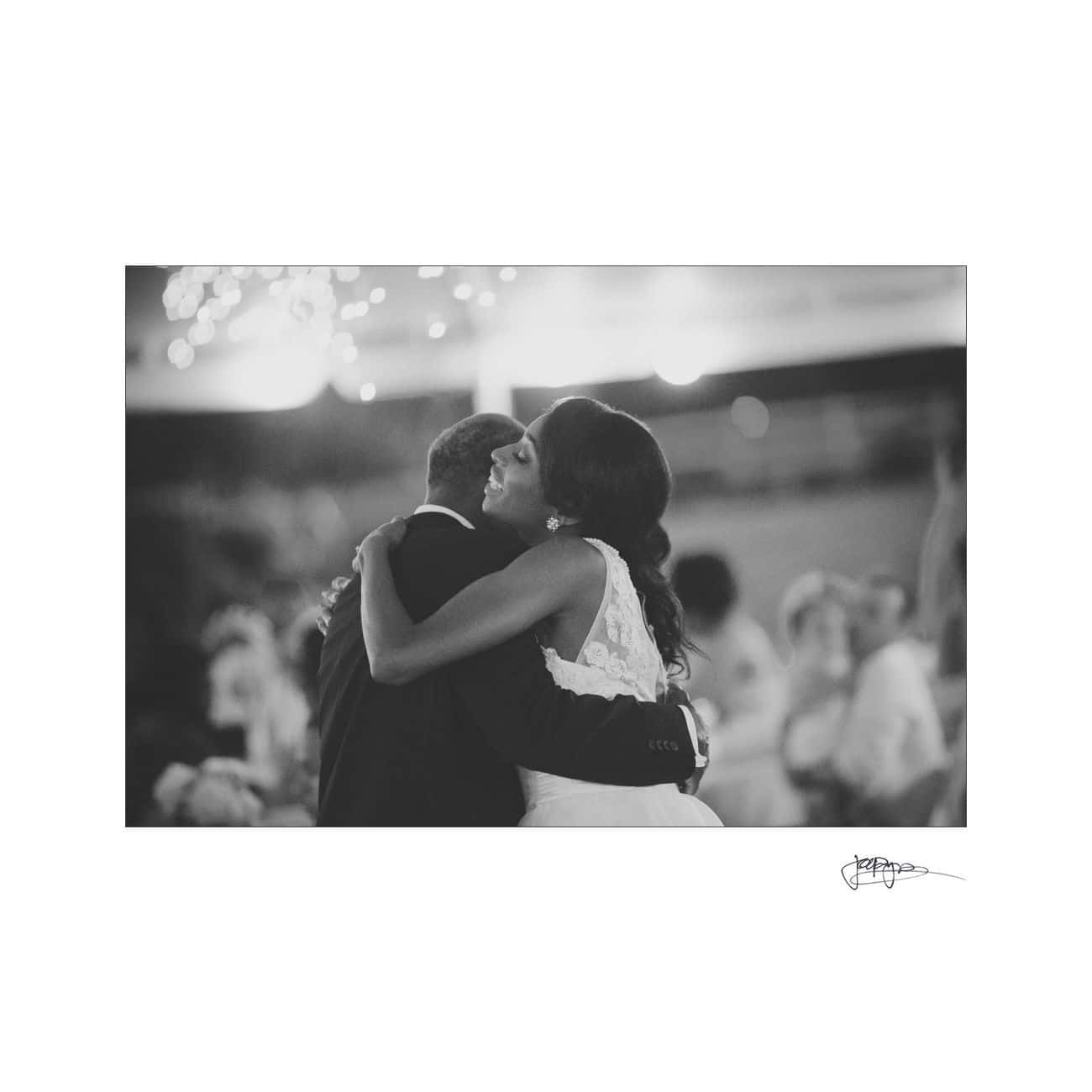A bride and groom share a tender embrace during their wedding dance. The image is in black and white, capturing the joyful moment with blurred guests in the background and soft lights overhead.