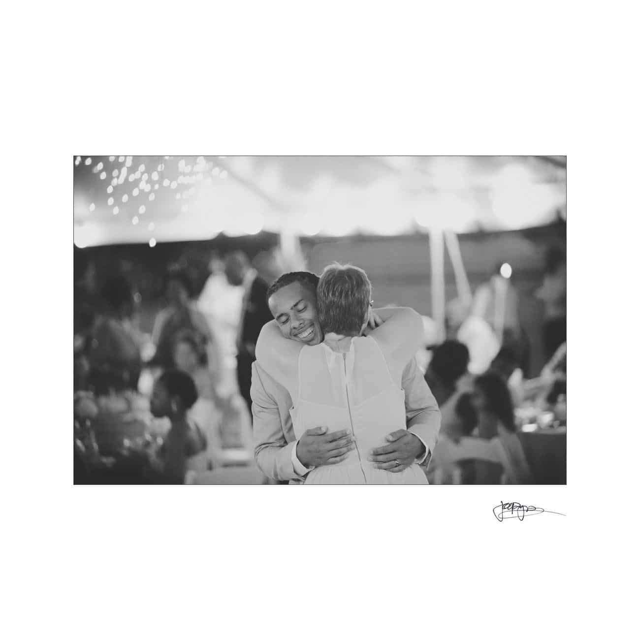 A black-and-white photo of two people hugging warmly at a festive gathering, surrounded by softly lit string lights and blurred figures in the background.