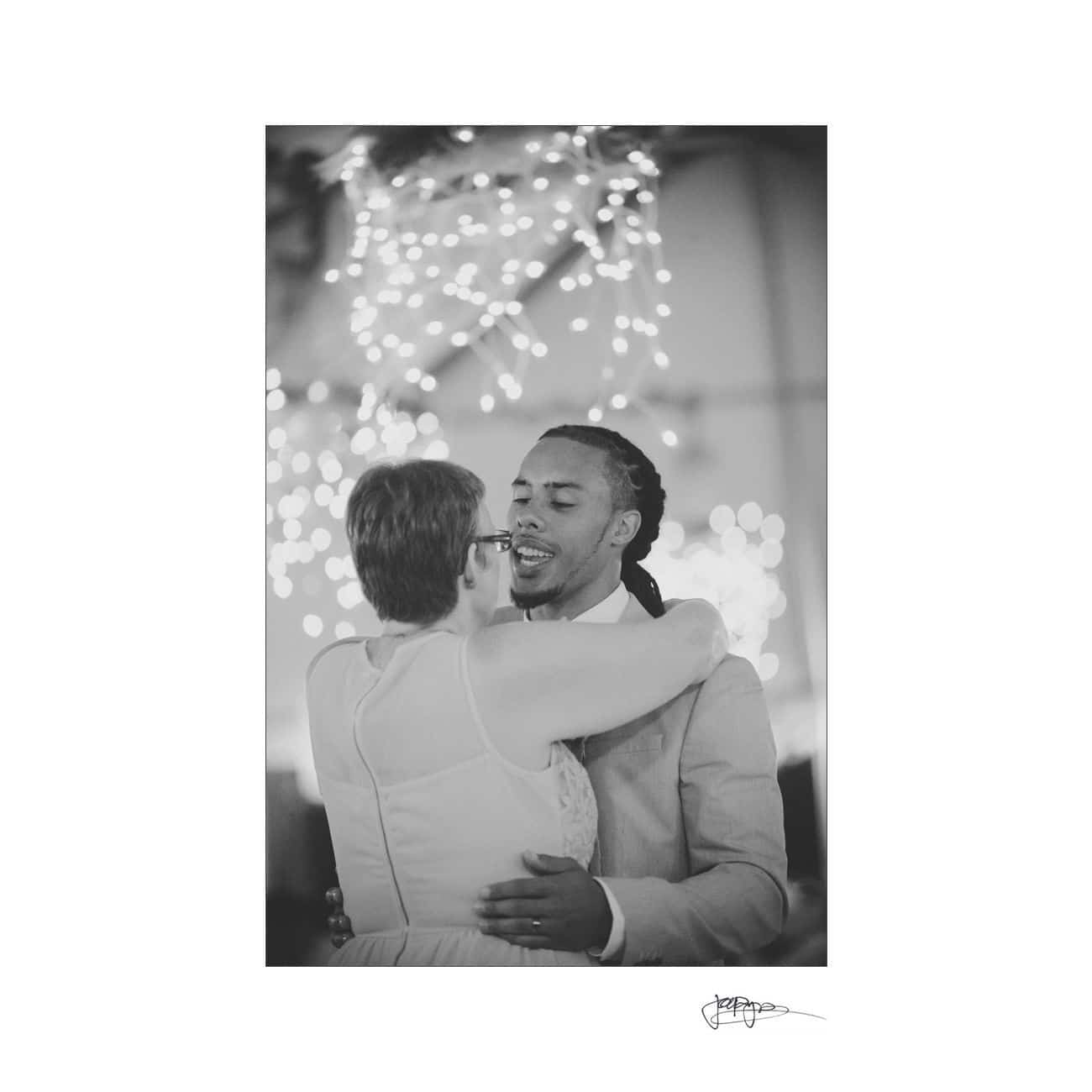 A black and white image of a couple dancing closely. They are embraced, looking at each other with smiles. The background is softly lit with string lights, creating a warm and intimate atmosphere.