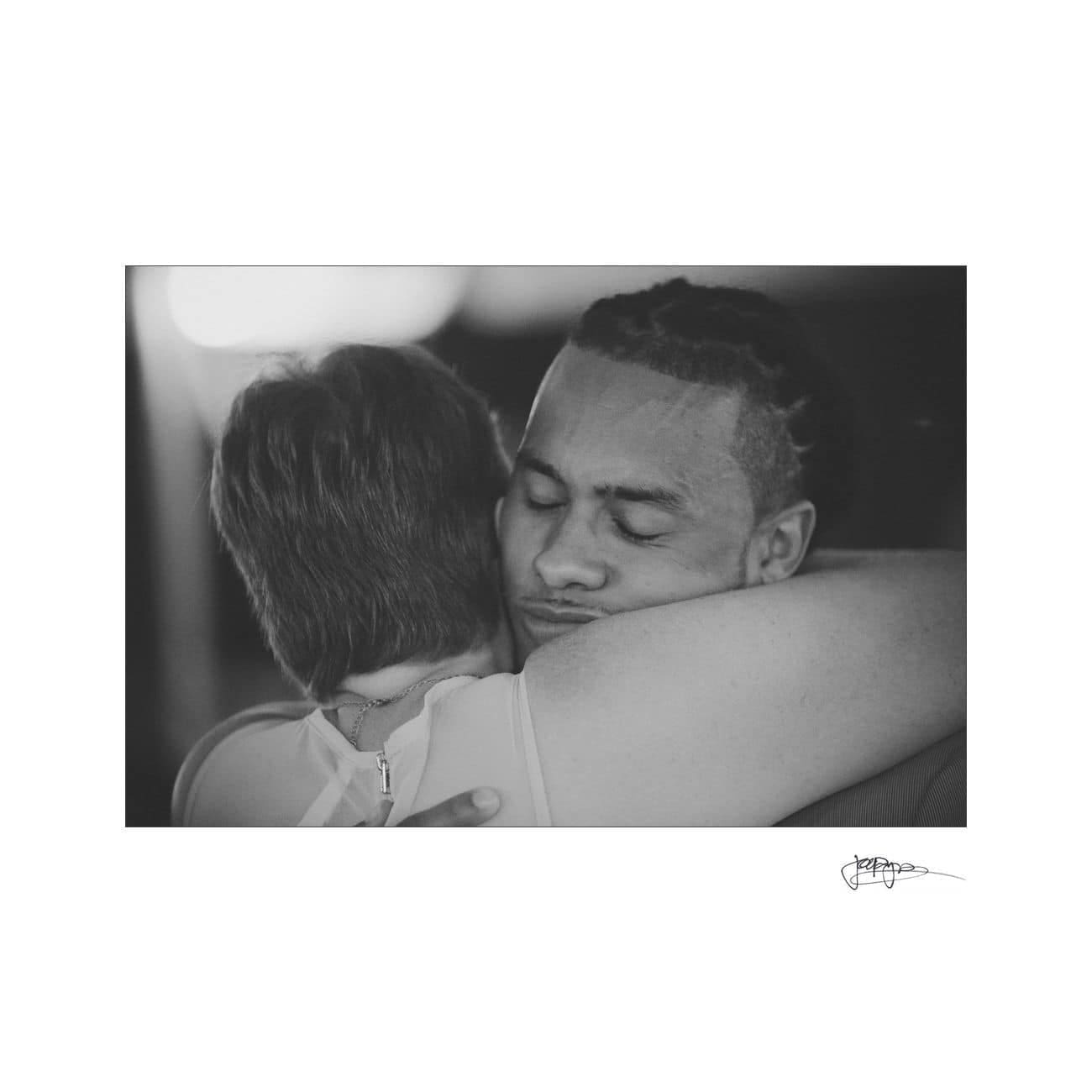 A black and white photo of a man and woman embracing tightly. The womans back is to the camera, while the man faces the camera with his eyes closed, conveying a sense of comfort and peace.