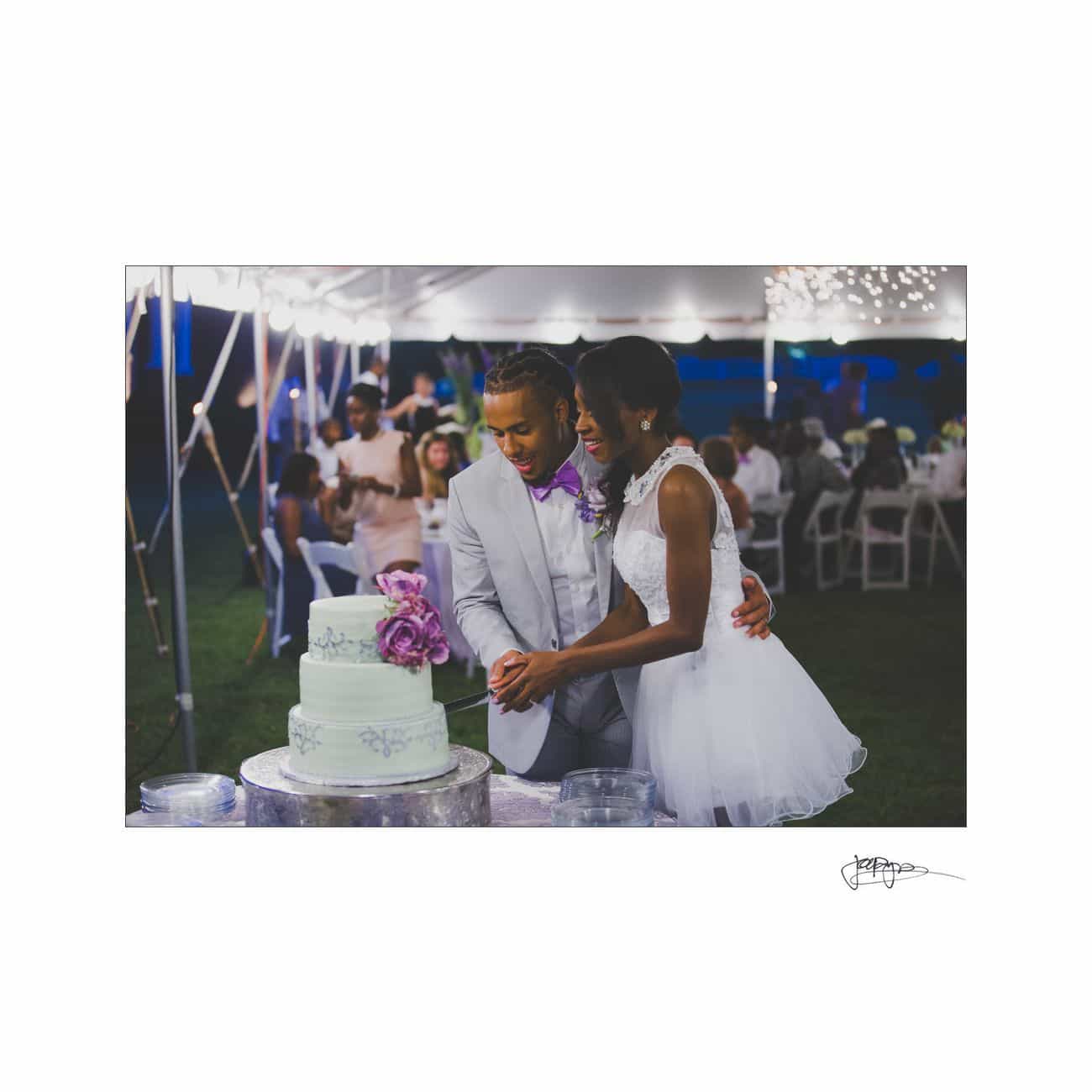 A couple in wedding attire is cutting a tiered cake with purple flowers under a tent. Guests are seated at tables in the background, and string lights are hanging above, creating a festive atmosphere.