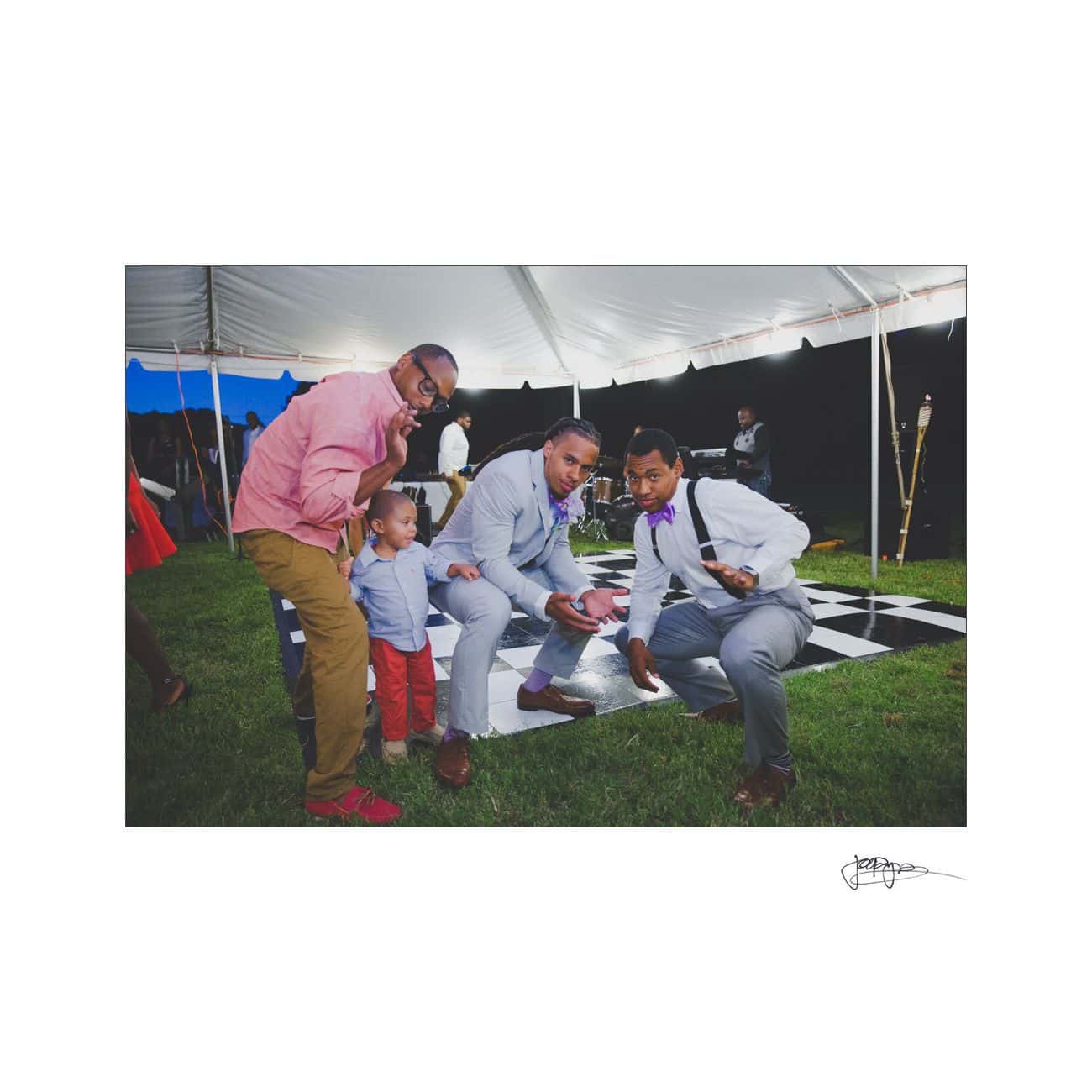 Four people, including a small child, pose playfully under a tent at an outdoor event. They are dressed in semi-formal attire, standing on grass with a checkerboard mat nearby. The atmosphere is casual and joyful.
