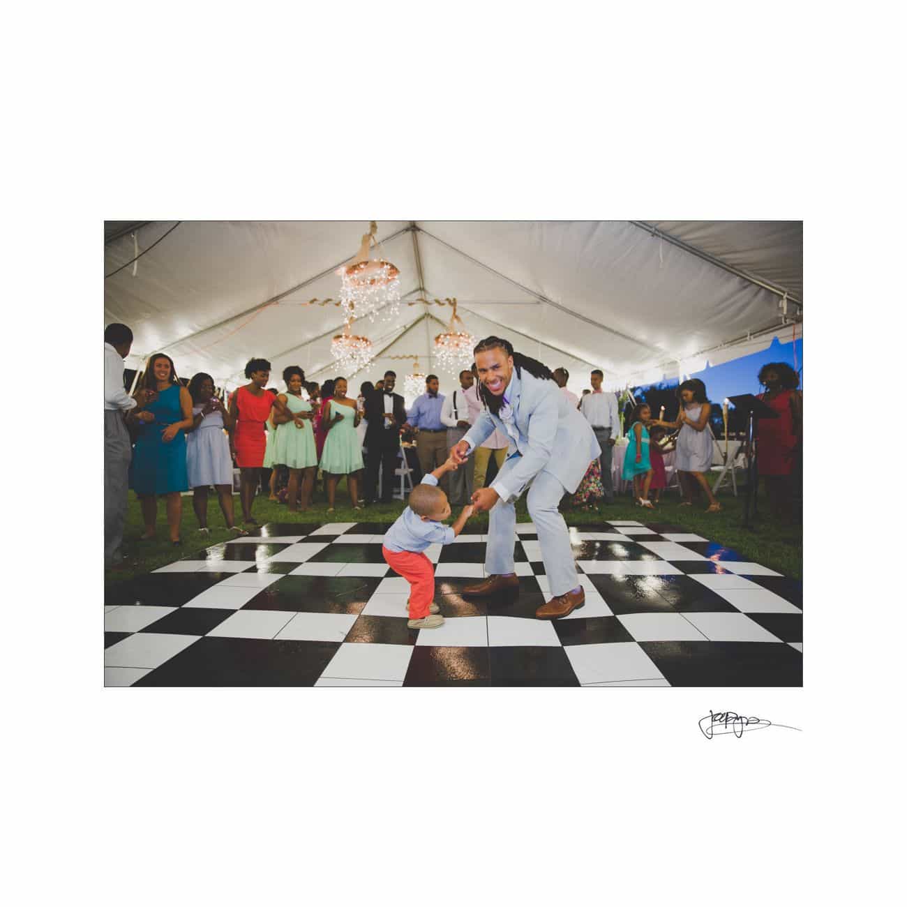 A man in a suit dances joyfully with a young child on a black-and-white checkered floor. They are surrounded by a crowd under a large white tent, with a chandelier above them.