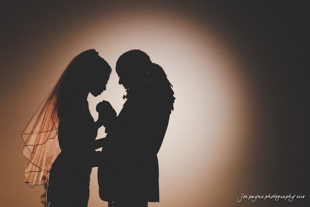 Silhouette of a bride and groom facing each other, holding hands tenderly, with a soft, glowing light behind them. Captured by Chapel Hill Wedding Photography, the couple shares an intimate moment as the bride’s veil gently frames their connection.