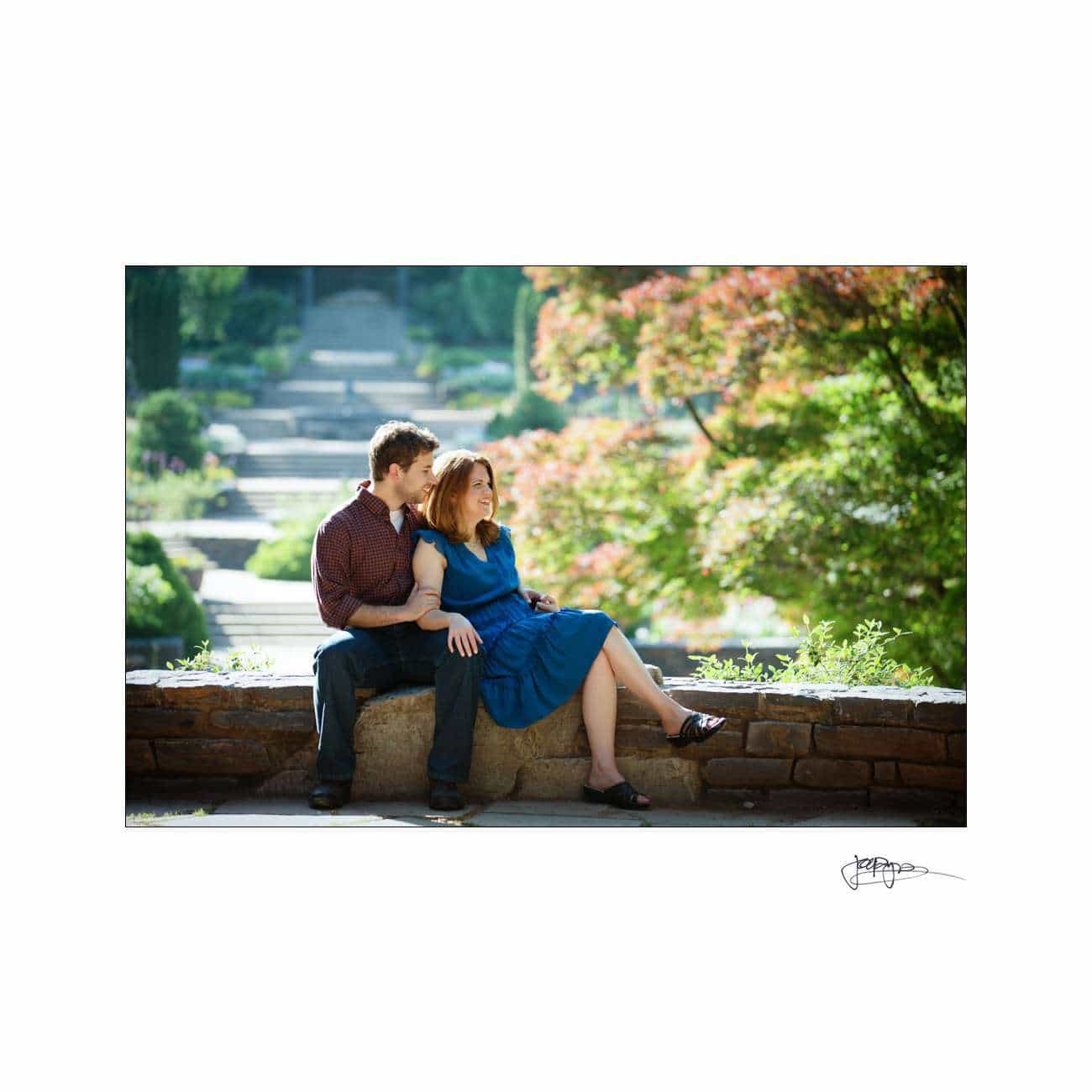 A couple sits on a stone wall in a sunlit park. The man, wearing a red shirt, sits beside the woman in a blue dress. They appear relaxed and content as they enjoy the lush greenery and vivid colors of the trees around them.