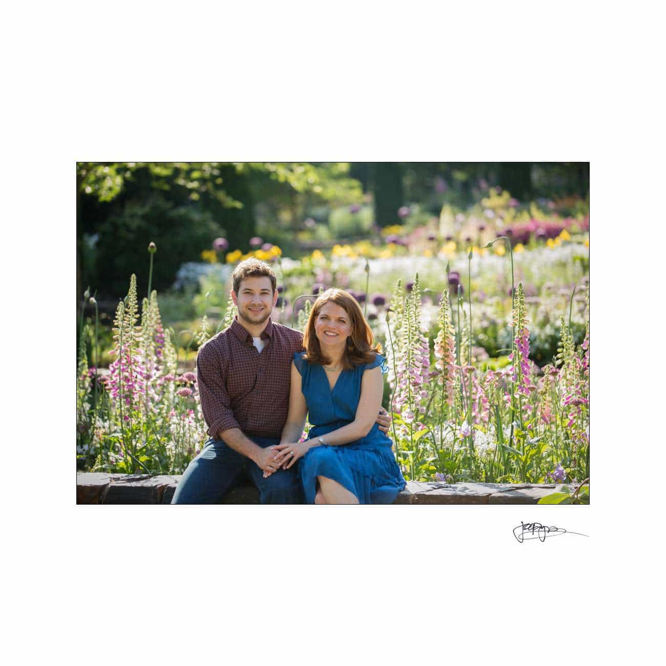 A couple sits on a low stone wall in a blooming garden, surrounded by vibrant purple and pink flowers. They hold hands and smile at the camera, with sunlight filtering through the lush greenery.