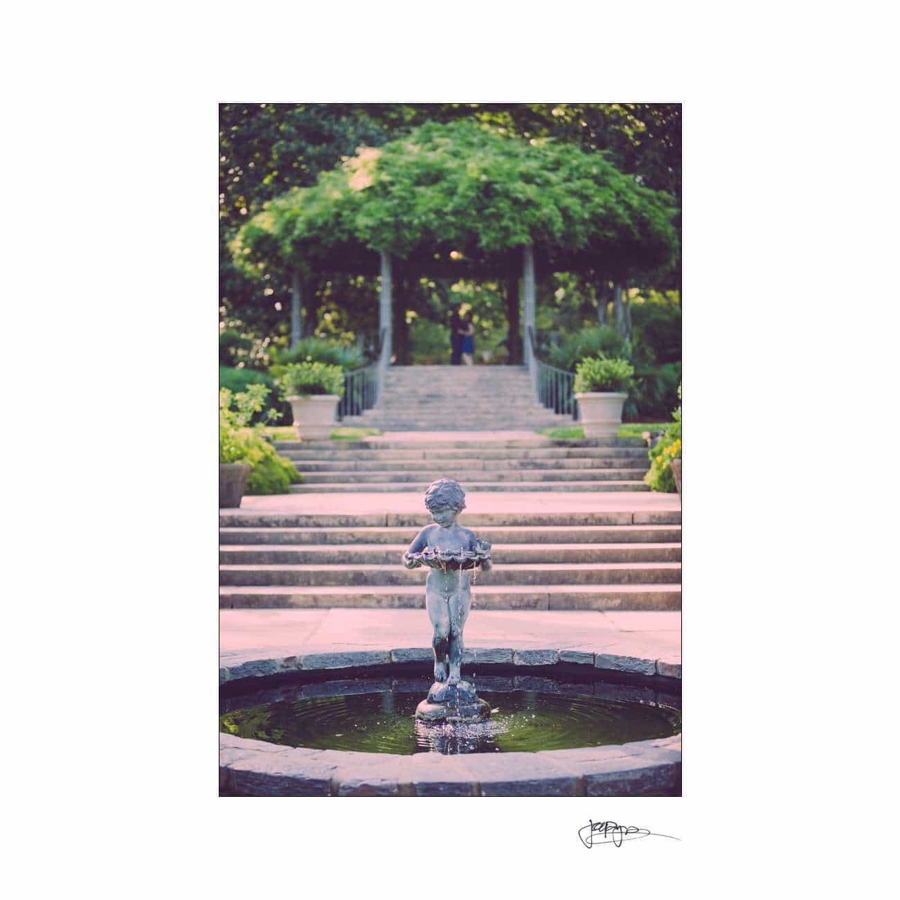 A serene garden scene with a small statue of a child at the center of a circular fountain. In the background, stone steps lead up to a tree-covered archway. Green foliage and decorative planters line the path.