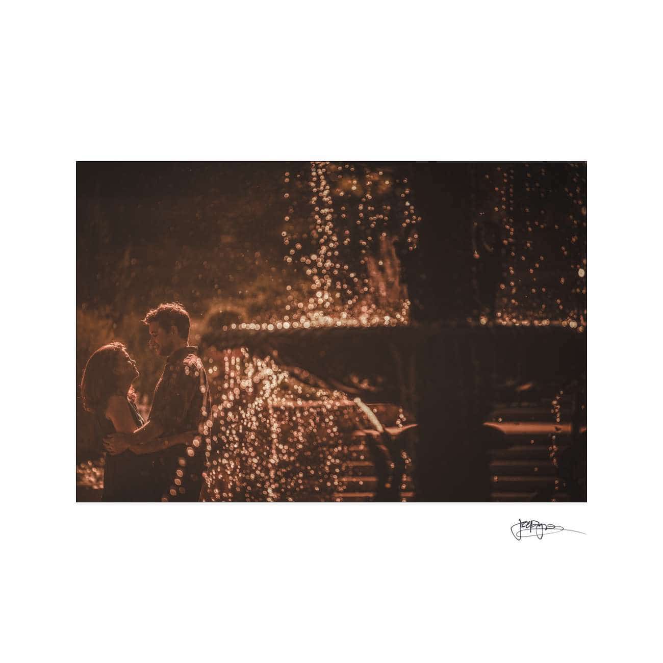 A couple stands close together, facing each other amidst sparkling water droplets in a softly lit, romantic setting. The warm light creates a dreamy atmosphere around them.