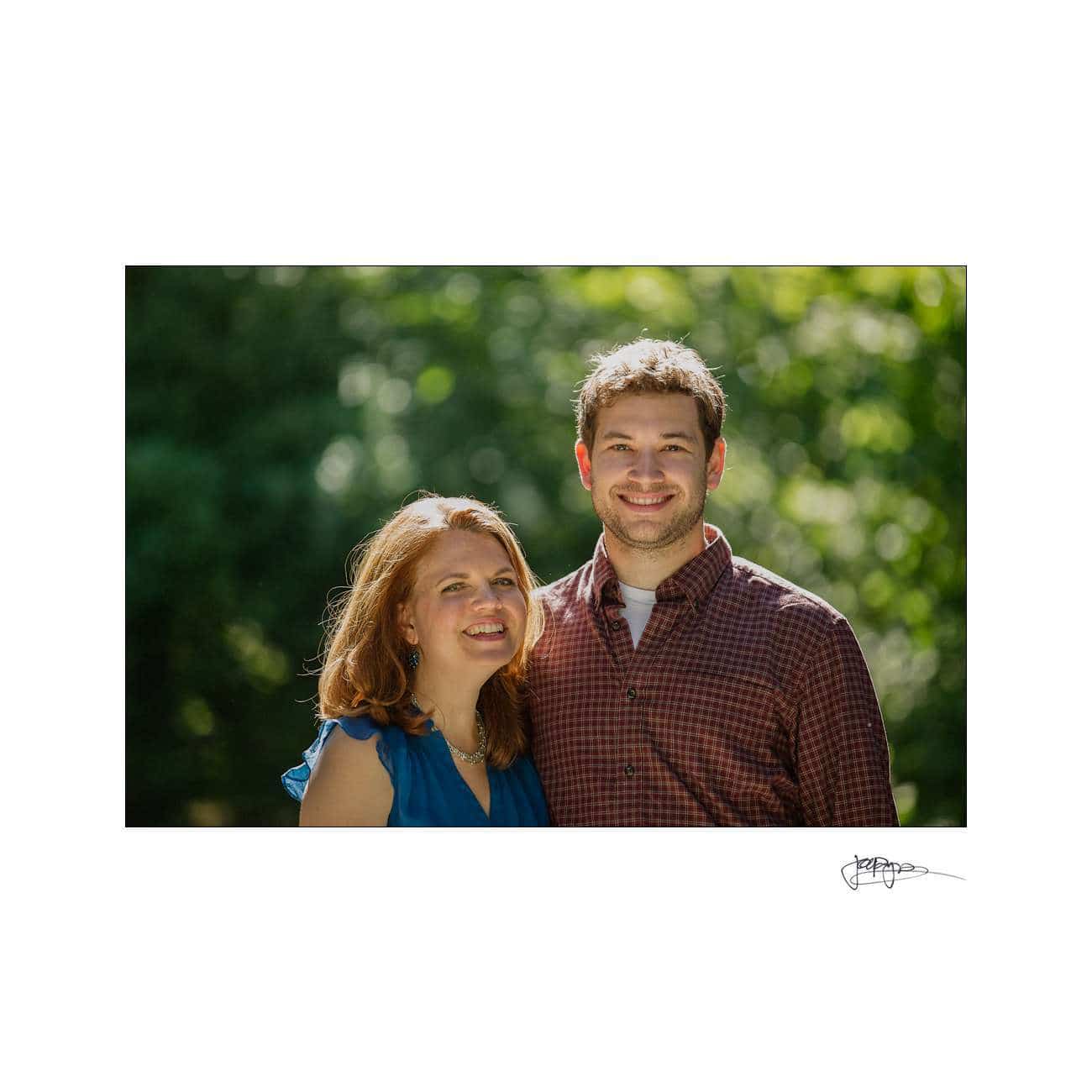 A smiling woman in a blue dress stands next to a smiling man in a red checkered shirt outdoors, with green foliage softly blurred in the background during their Duke Gardens engagement session.