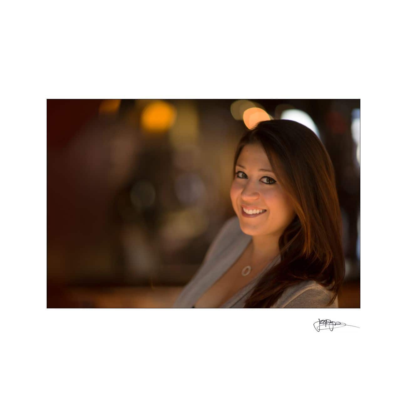 A woman with long hair smiles at the camera. She is wearing a gray top and a necklace with a circular pendant. The background is softly blurred with warm tones.
