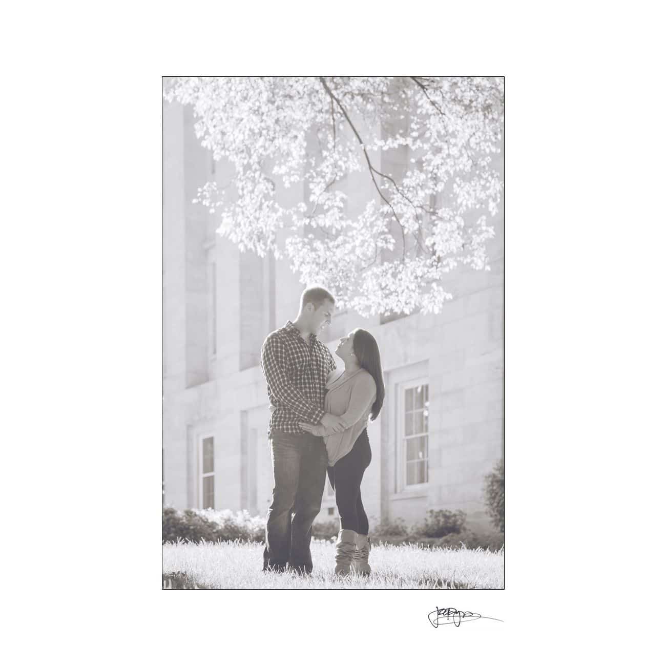 A sepia-toned image shows a couple standing on grass, facing each other under a tree with sunlight filtering through the leaves. They hold hands and smile warmly at each other, with a building in the background.