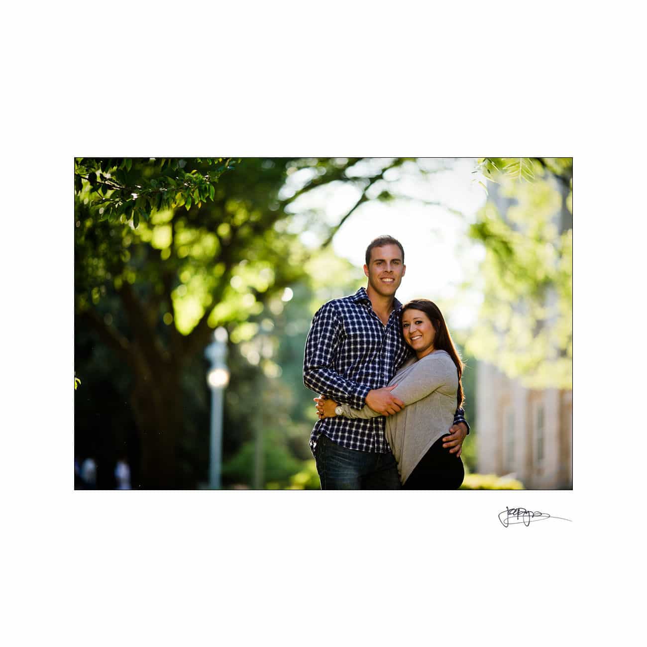 A couple embraces and smiles under sunlit trees on a bright day. The man wears a checkered shirt, and the woman wears a gray sweater. The background features greenery and a blurred building.