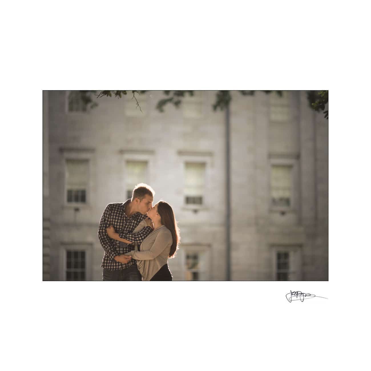 A couple embraces and leans in for a kiss in front of a large building with numerous windows. The scene is warmly lit, suggesting a romantic atmosphere.