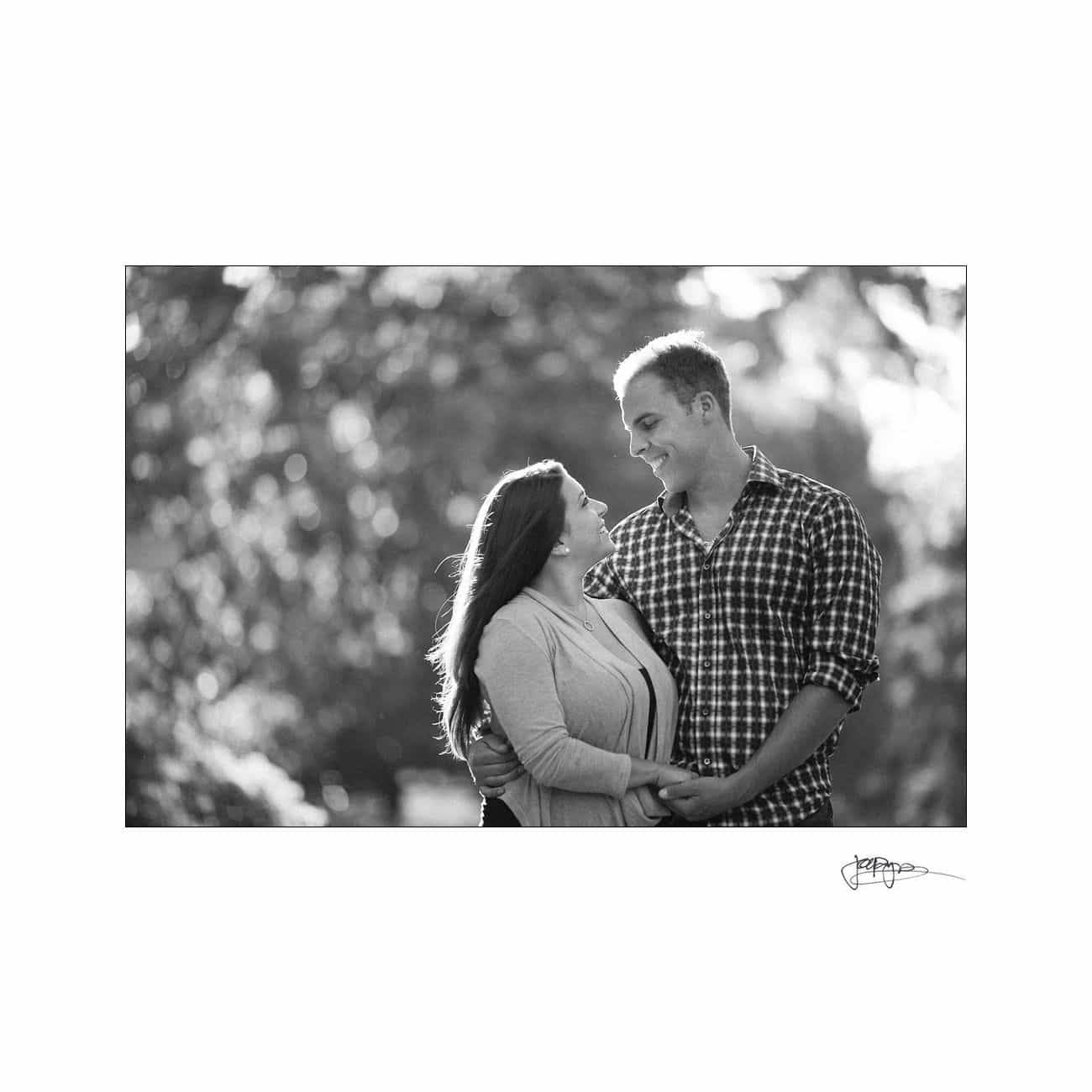 A black and white photo of a couple standing close together. They are outdoors, smiling at each other, with trees softly blurred in the background.