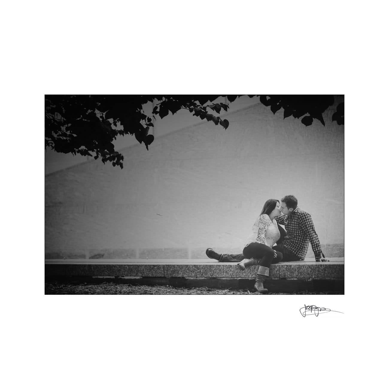 A black and white photo of a couple sitting together on a low stone wall, sharing an affectionate moment under a leafy tree. The background is a stark, textured wall, emphasizing the intimate scene in the foreground.