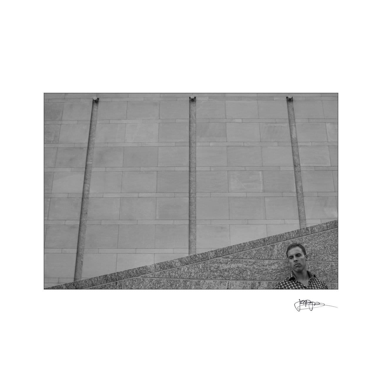 Black and white photo of a man standing on a sloped stone ledge, partially visible in the bottom right corner. The background features a large stone wall with vertical lines. A signature is in the bottom right corner.