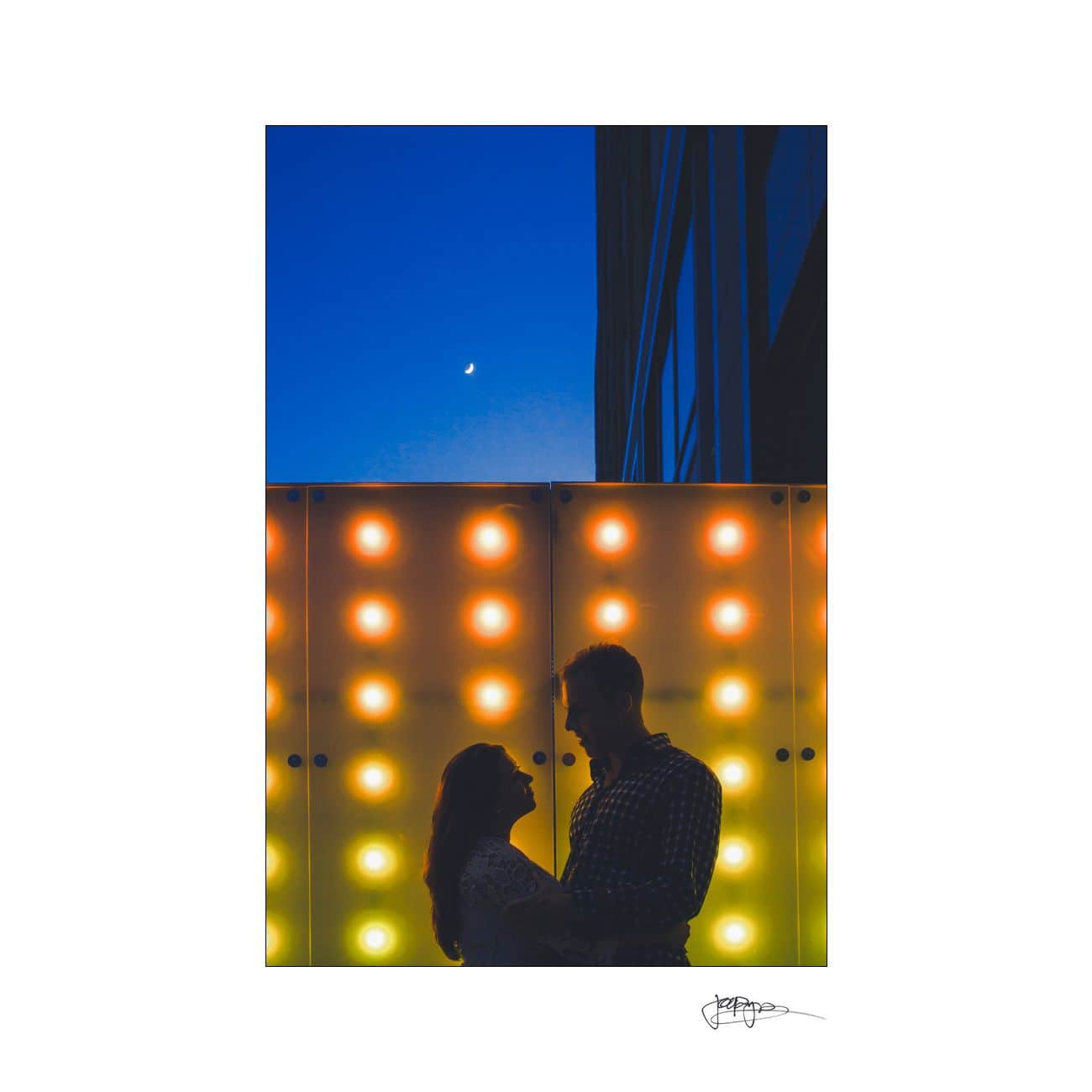 Silhouetted couple gazing at each other in front of a backdrop of colorful lights. A crescent moon is visible in the deep blue evening sky above.