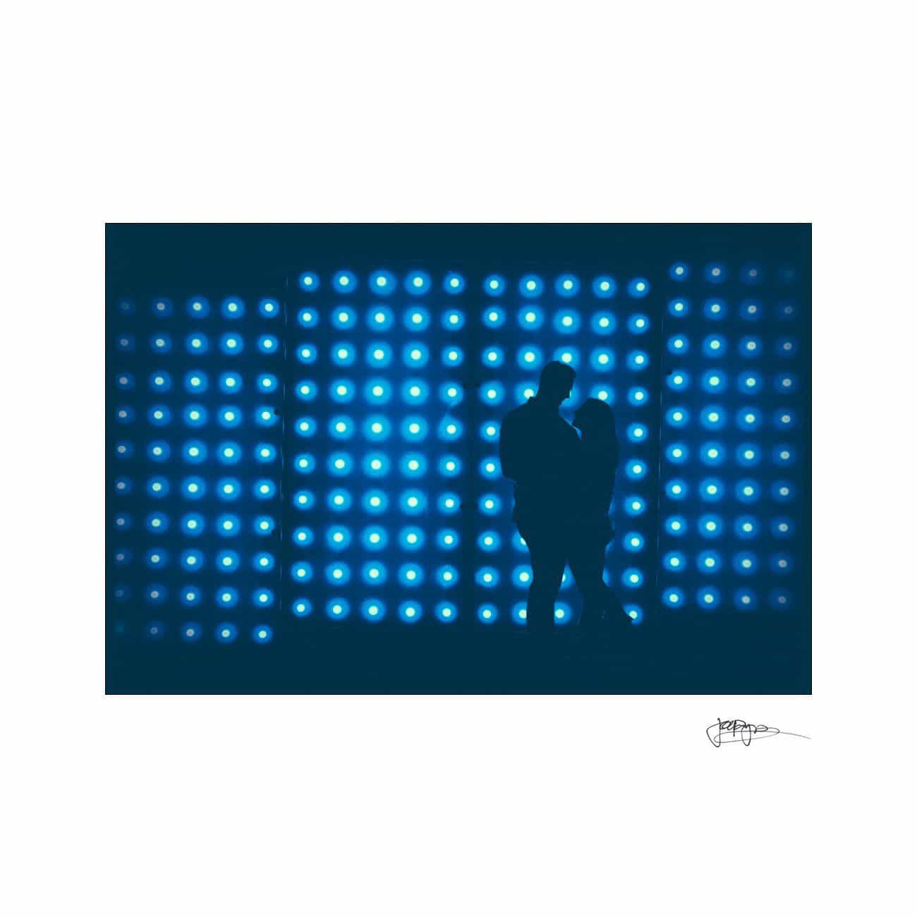 Silhouetted couple standing close together in front of a wall lit with an array of blue circular lights, creating a romantic and modern atmosphere.