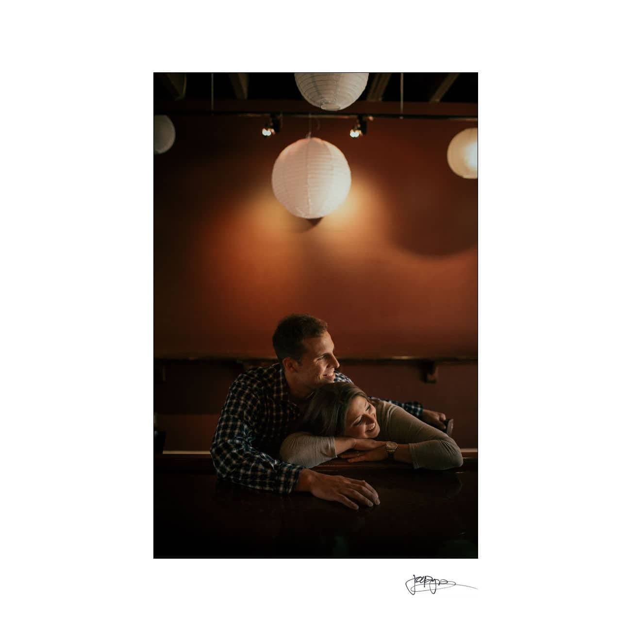 A couple leans on a bar counter, embracing under warm, soft lighting. The man in a plaid shirt rests his head on her, while the woman smiles gently. Two paper lanterns hang in the background, adding a cozy atmosphere.