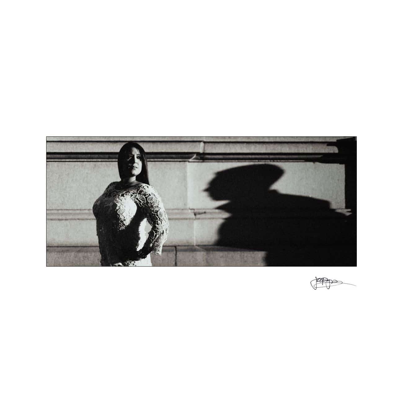 A woman in a lace dress stands confidently, casting a dramatic shadow on a textured wall. The shadow appears elongated and stylized, creating an intriguing contrast with the light. The image is signed by the photographer in the lower right corner.