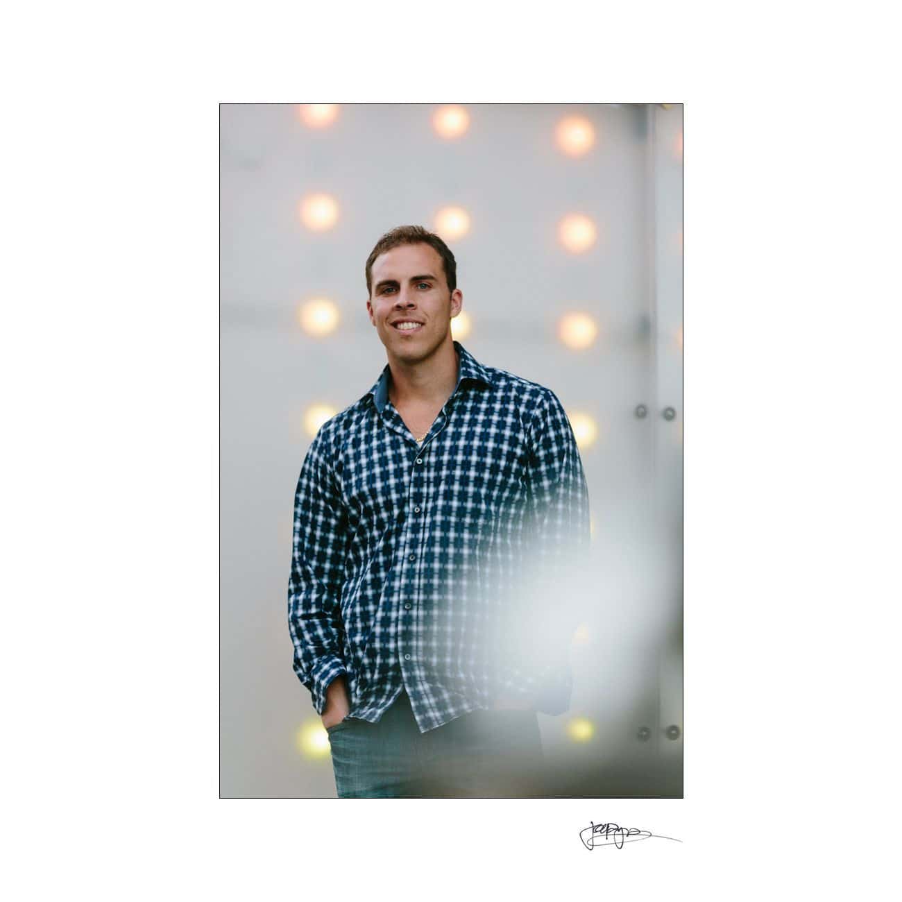 A man stands smiling, wearing a blue plaid shirt and jeans. The background features a soft bokeh effect with warm circular lights, creating a relaxed and casual atmosphere.