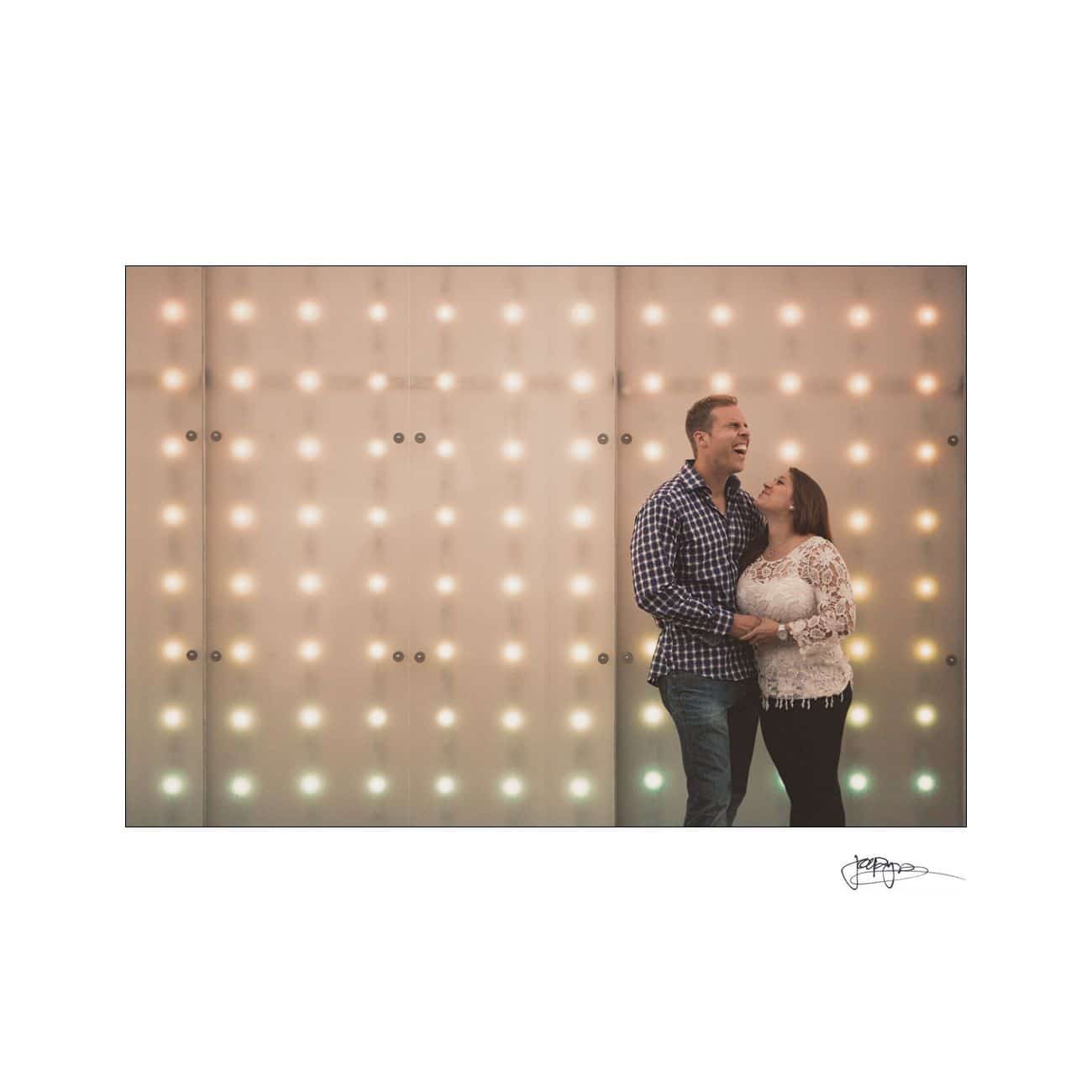 A couple stands laughing in front of a wall adorned with rows of warm, glowing lights. The man wears a checkered shirt and jeans, while the woman wears a lace top and jeans. The atmosphere is joyful and intimate.