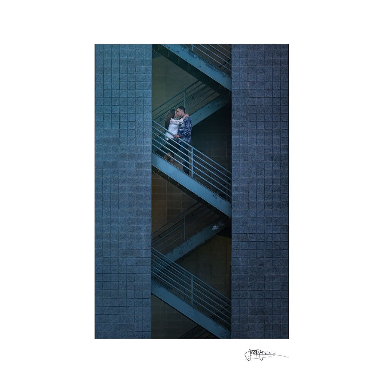 A couple embraces on a metal staircase between dark blue building walls. The structure creates a geometric pattern, emphasizing the vertical lines and contrasting the intimate moment with an industrial environment.