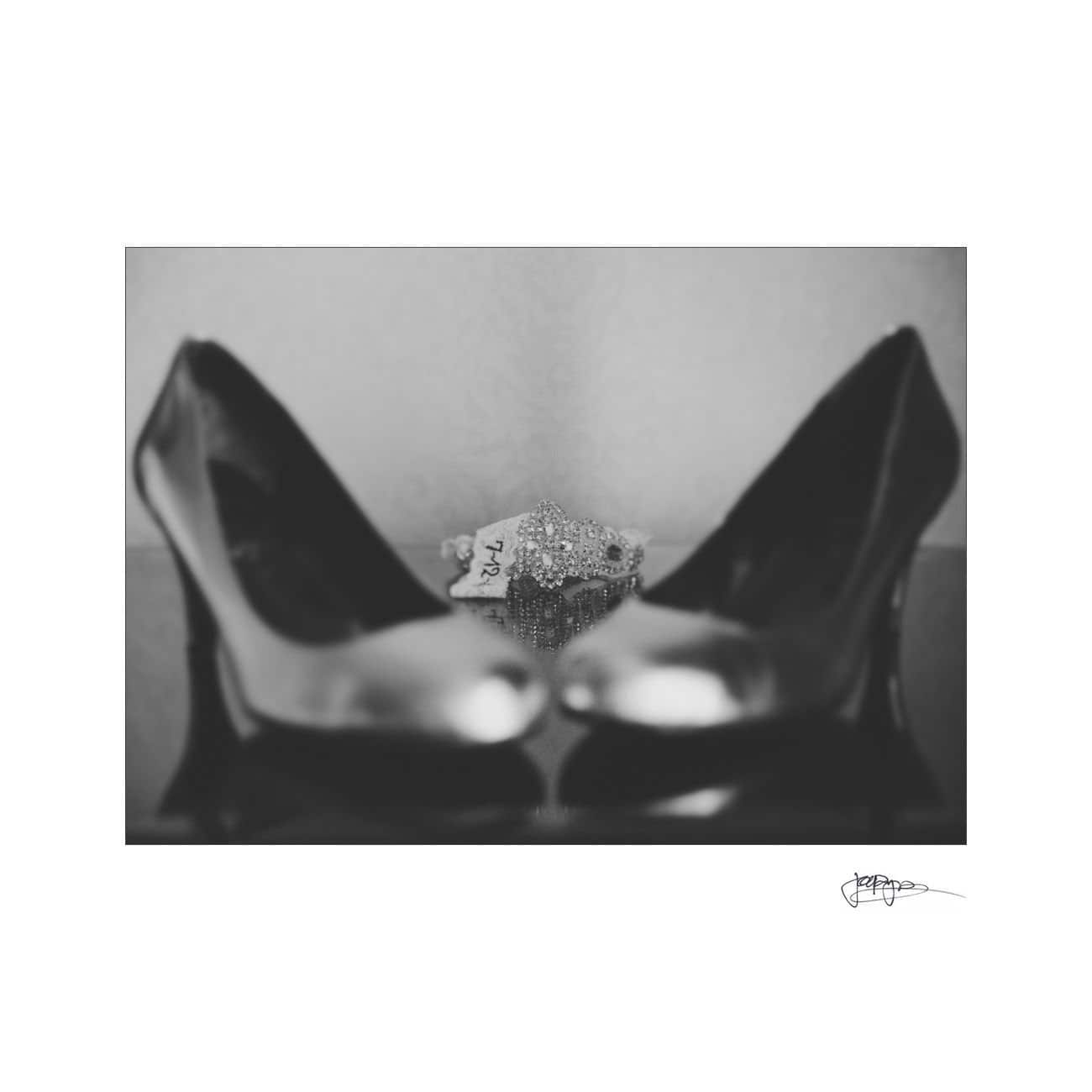Black and white image of a pair of shiny high heels in the foreground, with a sparkling jeweled tiara placed behind them. The background is blurred, creating a focus on the shoes and the tiara.