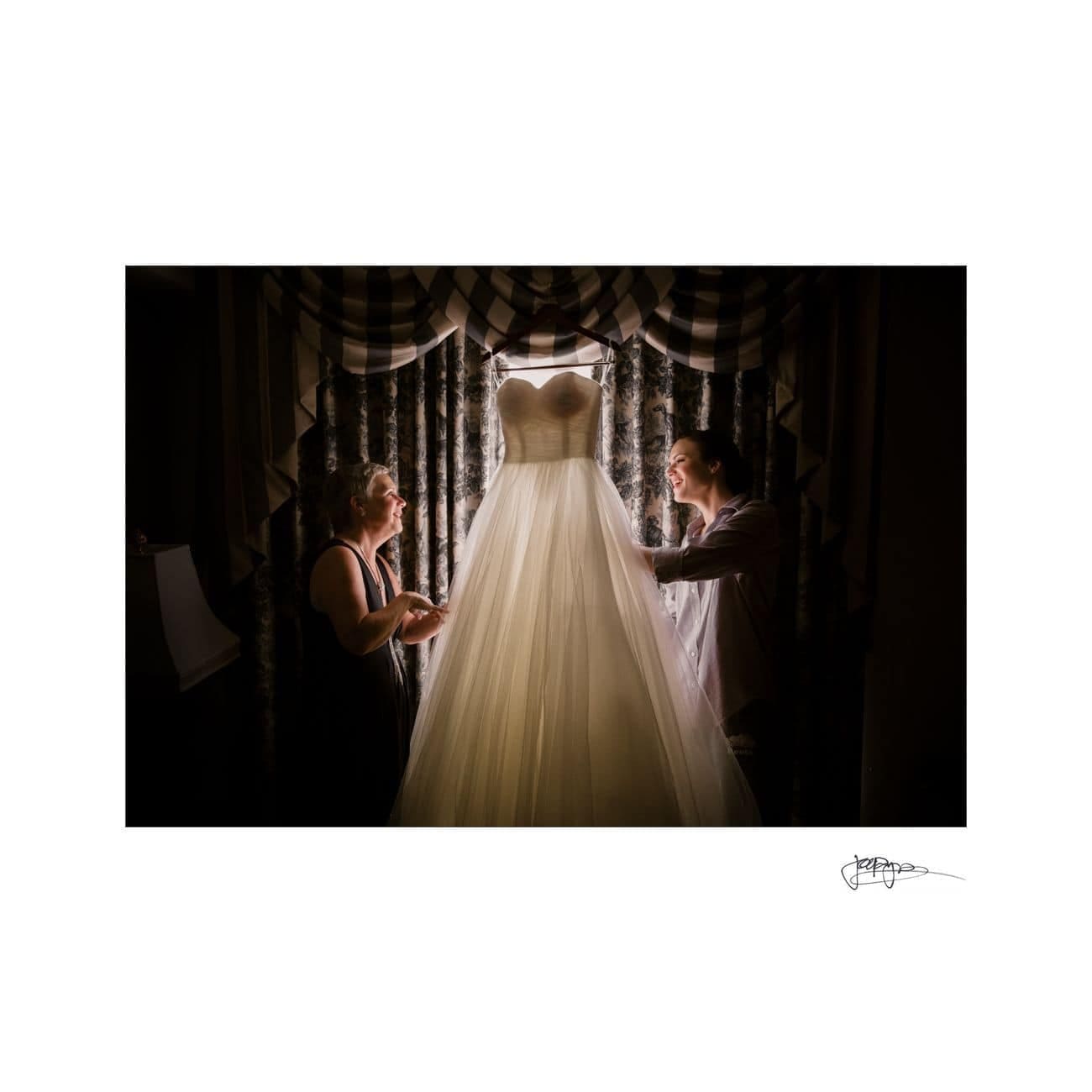 A bride and a woman admire a wedding dress hanging in front of a window, silhouetted against soft light. The dress is long and flowing, with a fitted bodice. Shadows and draped curtains create an intimate, serene atmosphere.