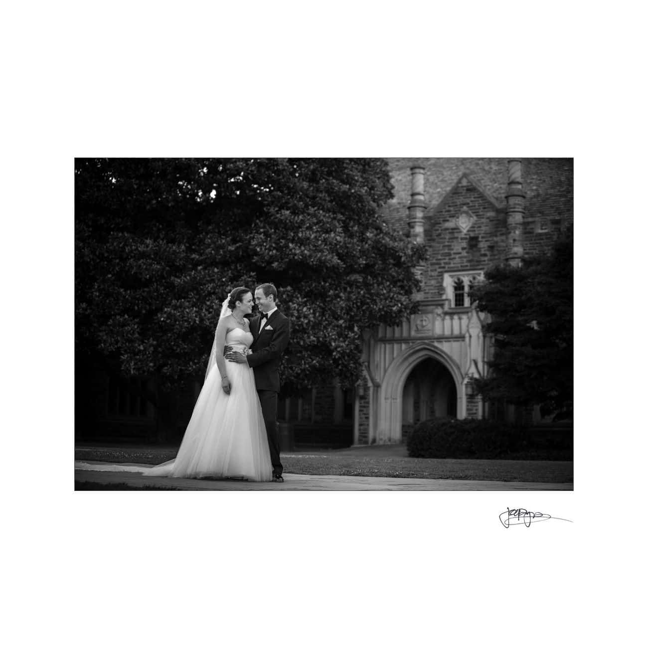 A bride and groom stand facing each other and holding hands, dressed in wedding attire, in front of the arched stonework and trees of Duke Chapel, captured in black and white.