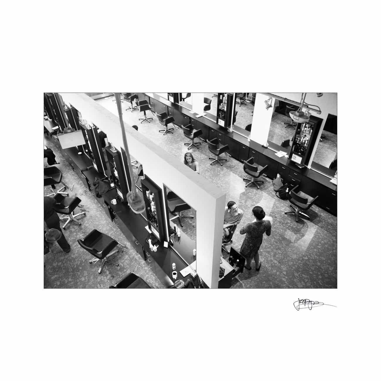 A black and white photo of a hair salon with multiple chairs and mirrors lining the walls. A stylist stands near a client seated in a chair. The salon has a modern, organized appearance with various hair products on the counters.