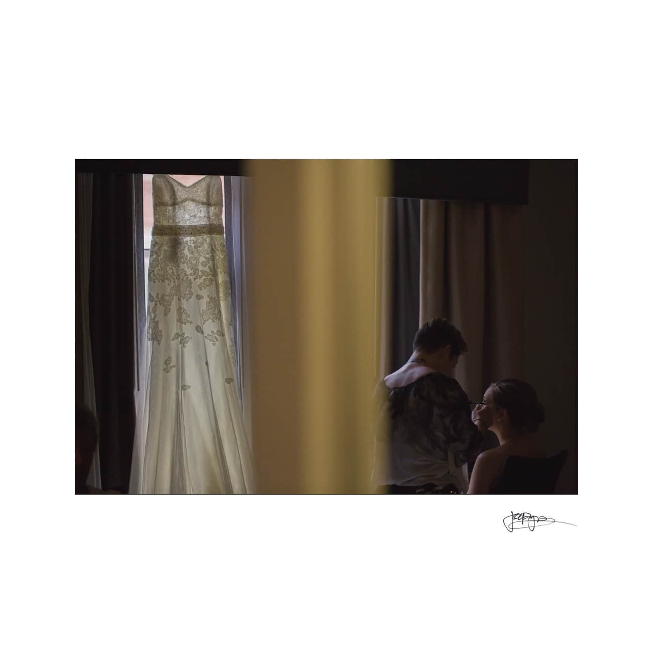 A wedding dress hangs in a window on the left side, with intricate lace details. On the right, a person is getting their makeup done in front of a mirror, partially obscured by dim lighting. The scene is calm and intimate.