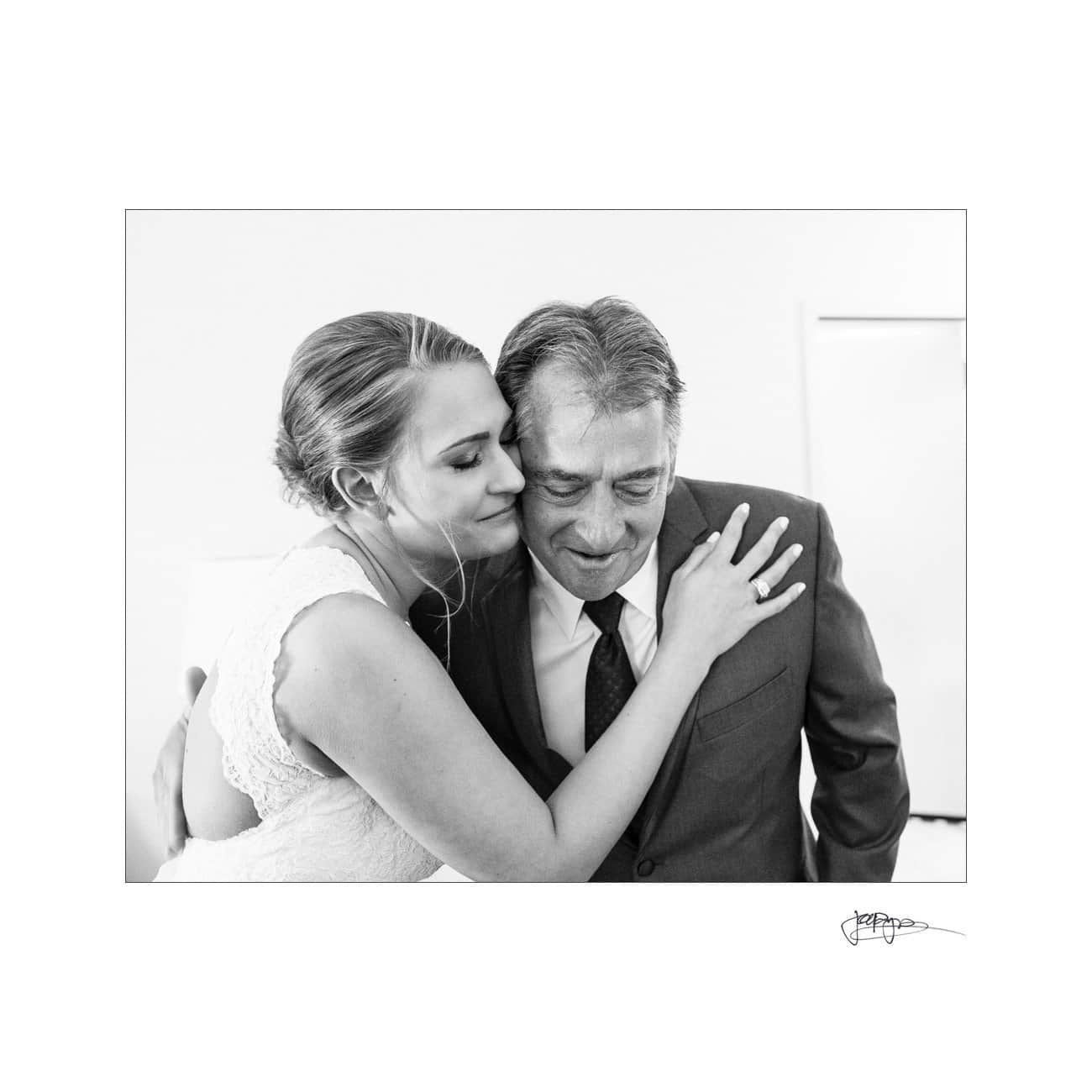 A bride in a white dress lovingly hugs an older man in a suit. They both have gentle smiles, sharing an emotional moment. The image is in black and white, enhancing the sentimental atmosphere.