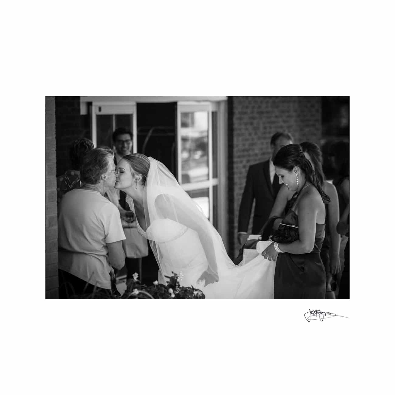 A bride in a veil kisses an older woman while others stand nearby. Another woman helps hold the brides dress. The scene is in black and white, adding a timeless feel to this intimate moment.