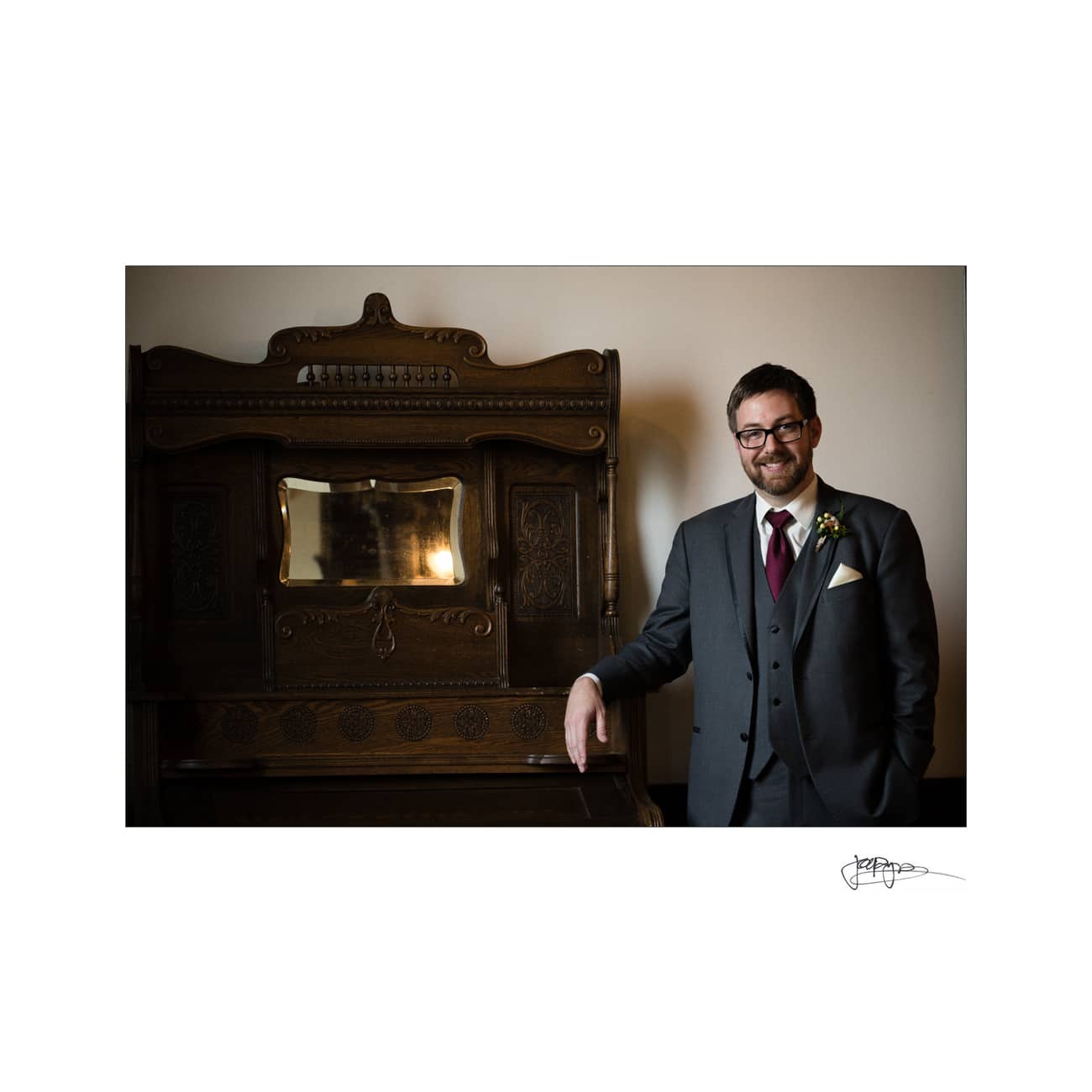 A man in a dark suit with glasses and a boutonniere leans casually against a vintage wooden cabinet with a mirrored panel. The setting is warmly lit, creating a cozy ambiance.