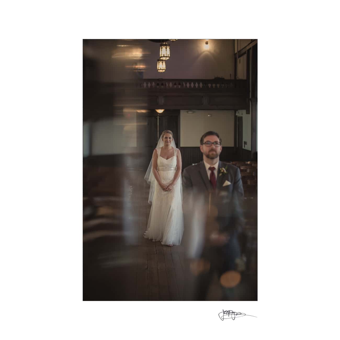 A bride in a white dress and veil stands in a softly lit, empty church. In the foreground, a blurred figure in a suit, possibly the groom, is visible. The atmosphere is serene and intimate.