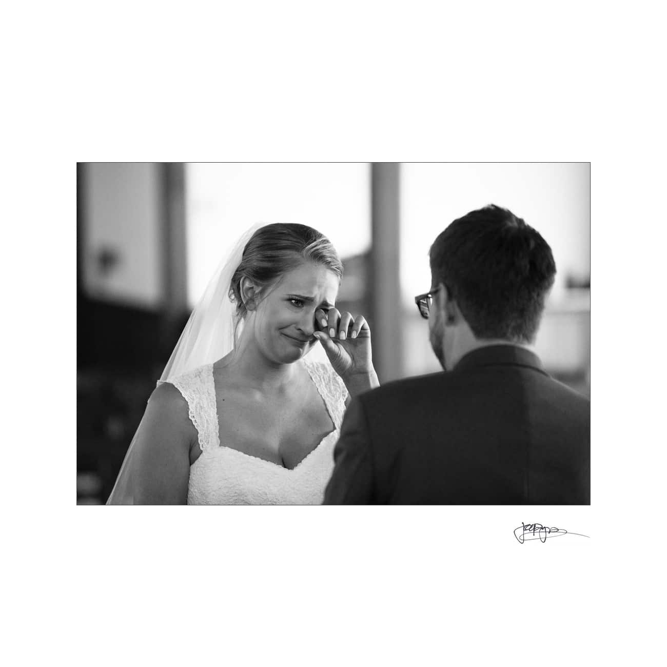 A bride in a wedding dress wipes a tear from her eye while facing the groom. The scene captures an emotional moment during the ceremony. The setting appears to be indoors, with soft lighting highlighting the couple.