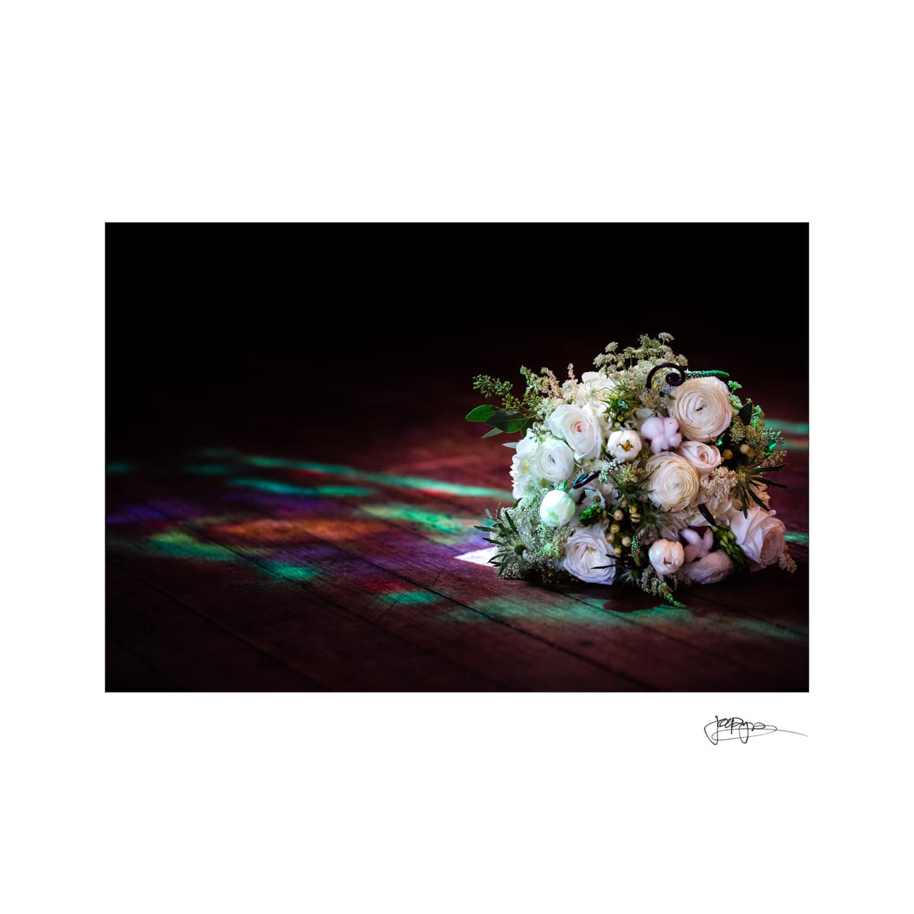 A bouquet of white roses and greenery is placed on a wooden floor. Colorful lights create a vibrant, patterned reflection beside it. The background is dark, adding contrast to the illuminated flowers.