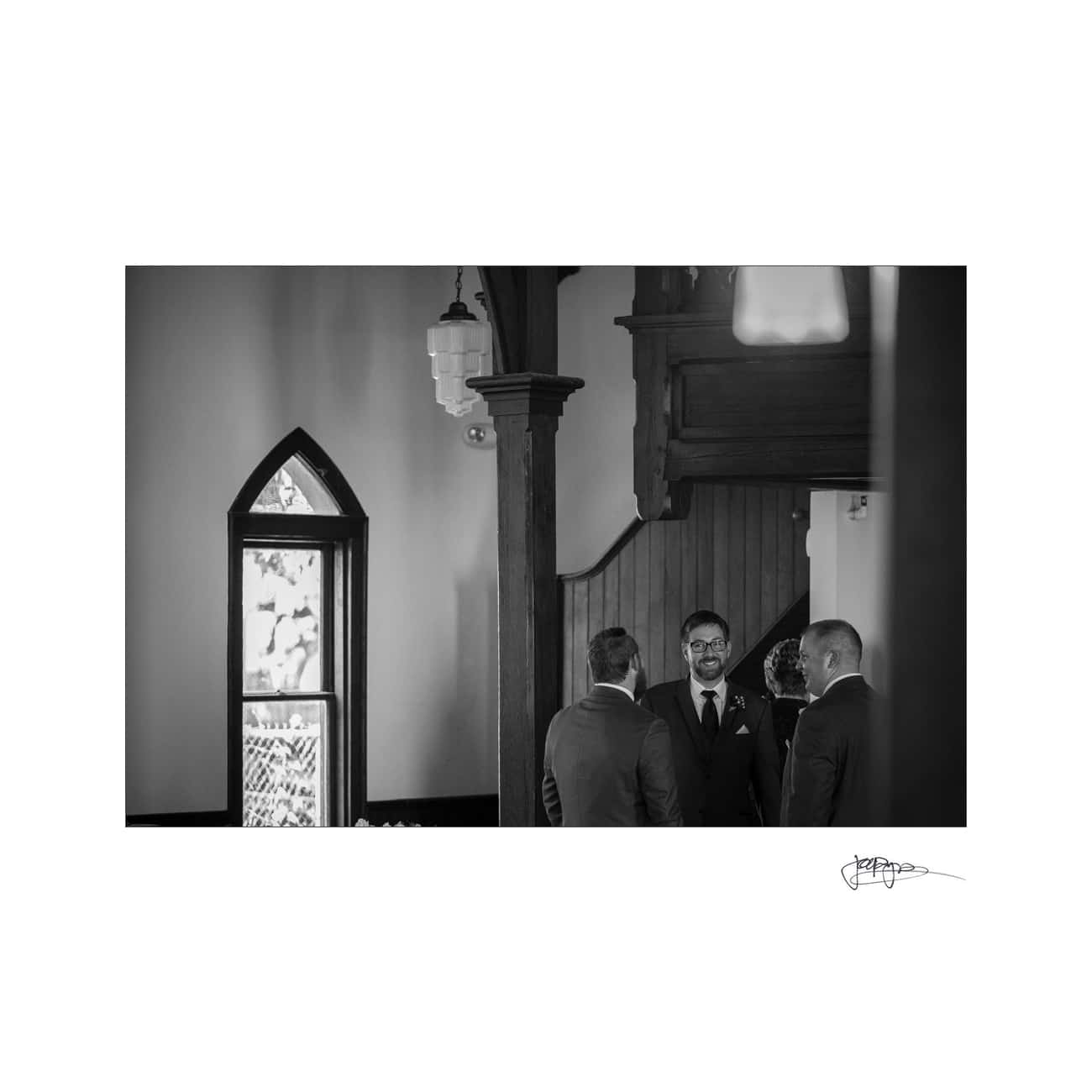 Black and white photo showing three men in suits standing and conversing inside a room with wooden details and a stained glass window. One man is smiling and looking directly ahead.