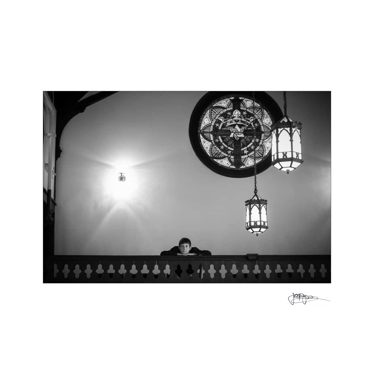 A person is sitting at a balcony, reading a book in a dimly lit room. Two hanging lamps illuminate the space, and a decorative stained glass window is visible in the background. The image is in black and white.