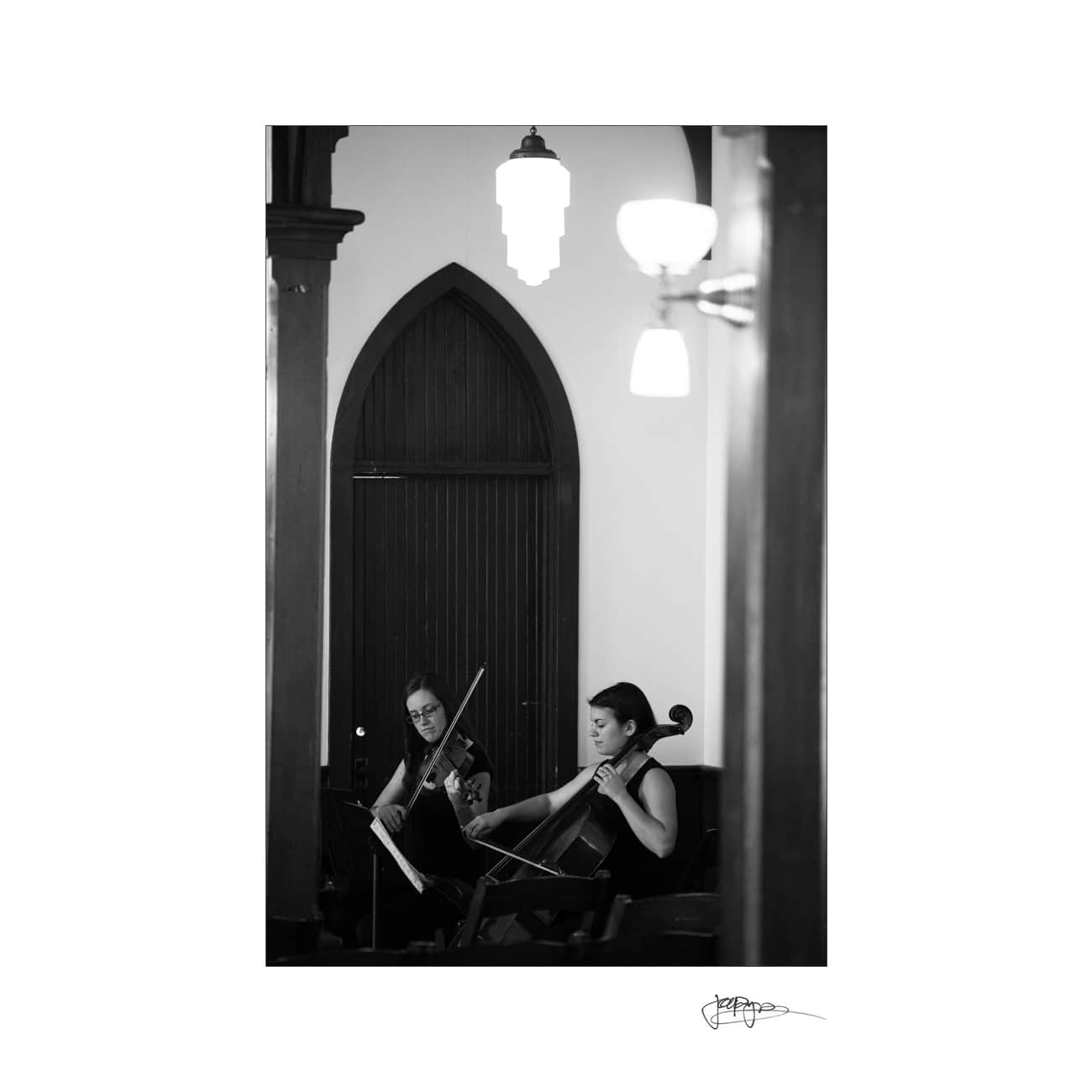 Black and white photo of two women playing string instruments in a room with arched wooden doors. One plays a violin, the other a cello. A pendant light casts a soft glow above them.