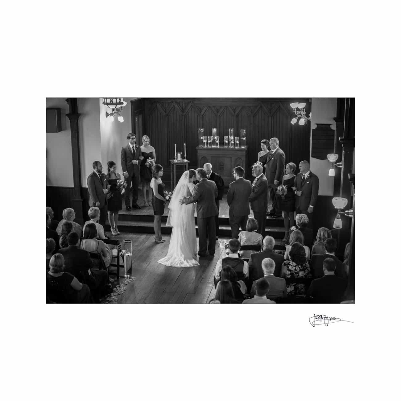 Black and white photo of a wedding ceremony in a church. The bride and groom stand facing the officiant, surrounded by the bridal party. Guests are seated, attentively watching. Candles are lit on the altar in the background.
