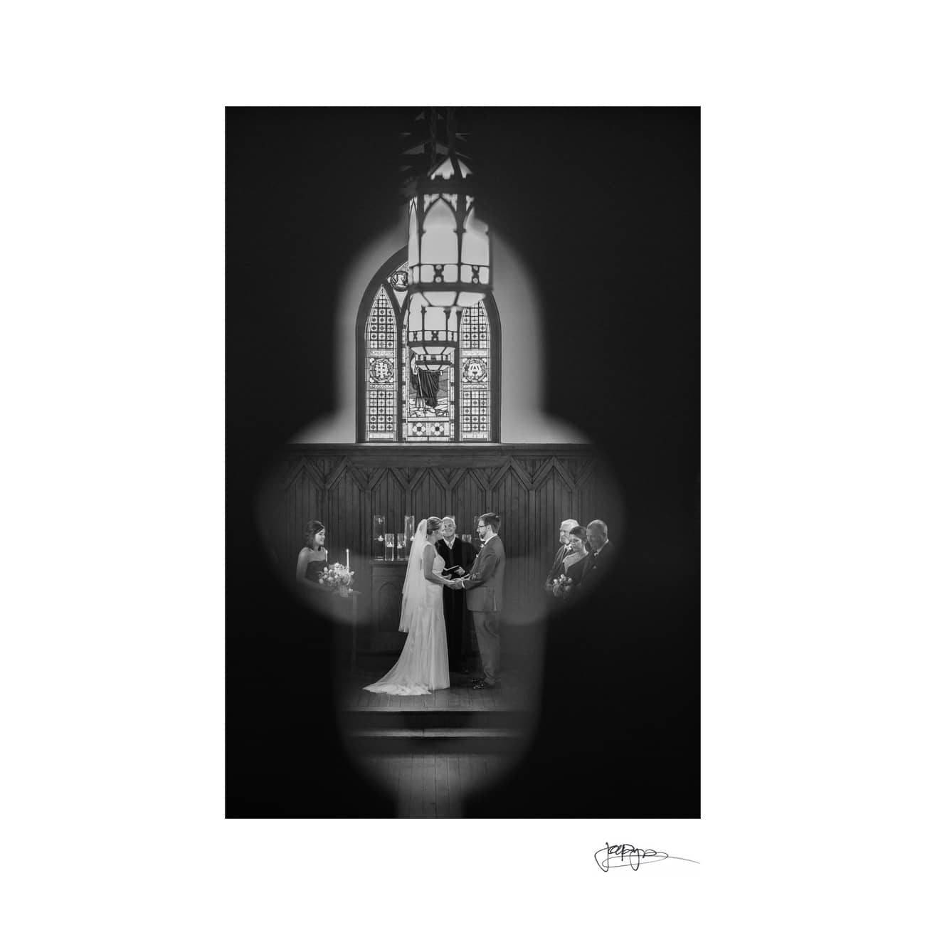 A black and white photo of a wedding ceremony viewed through a keyhole-shaped opening. The couple stands at the altar in front of a stained glass window, exchanging rings, with the officiant and guests in the background.