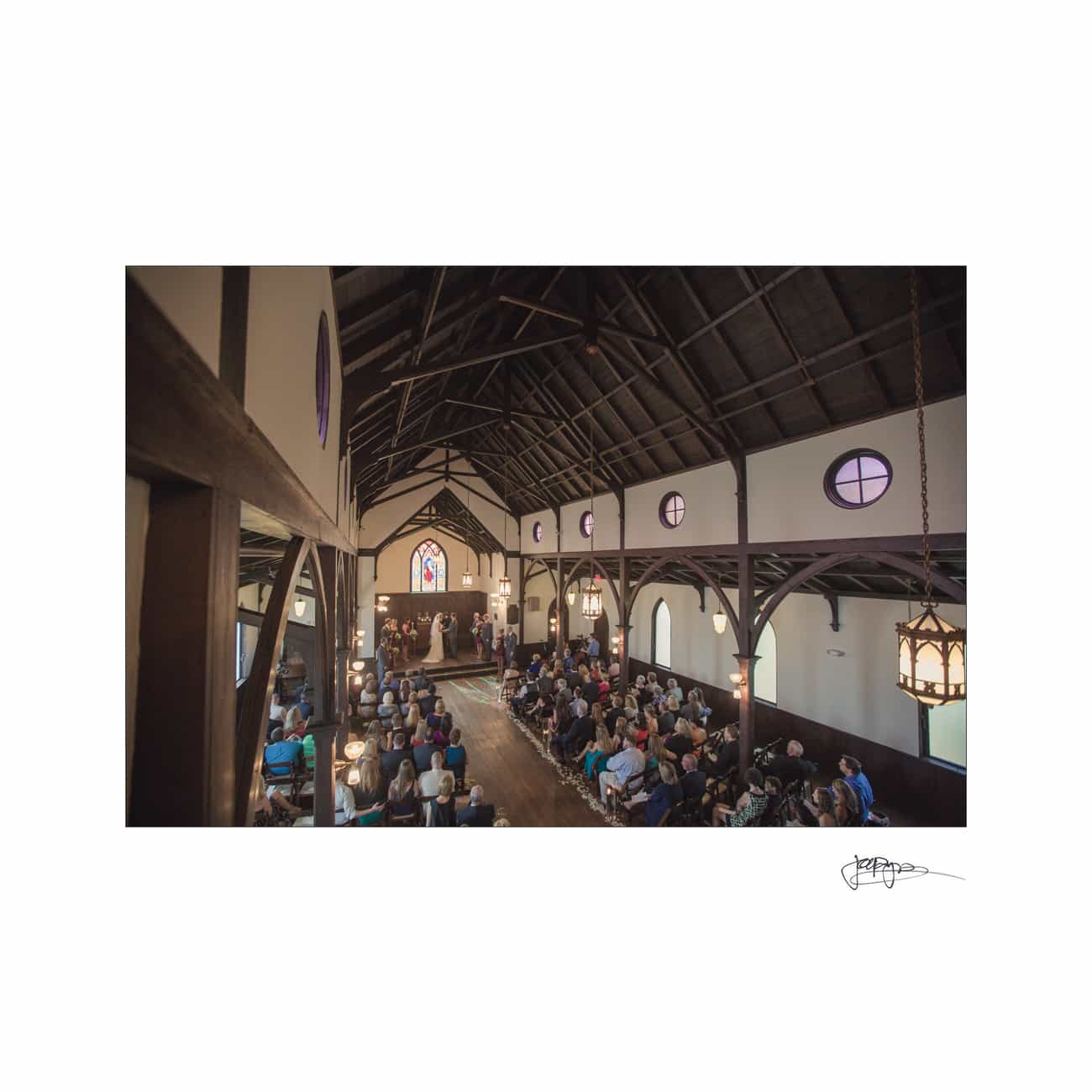 A wide-angle view of a church interior during a wedding ceremony. Guests are seated on both sides, facing the altar where the couple stands with the officiant. The church features wooden beams and stained glass windows.