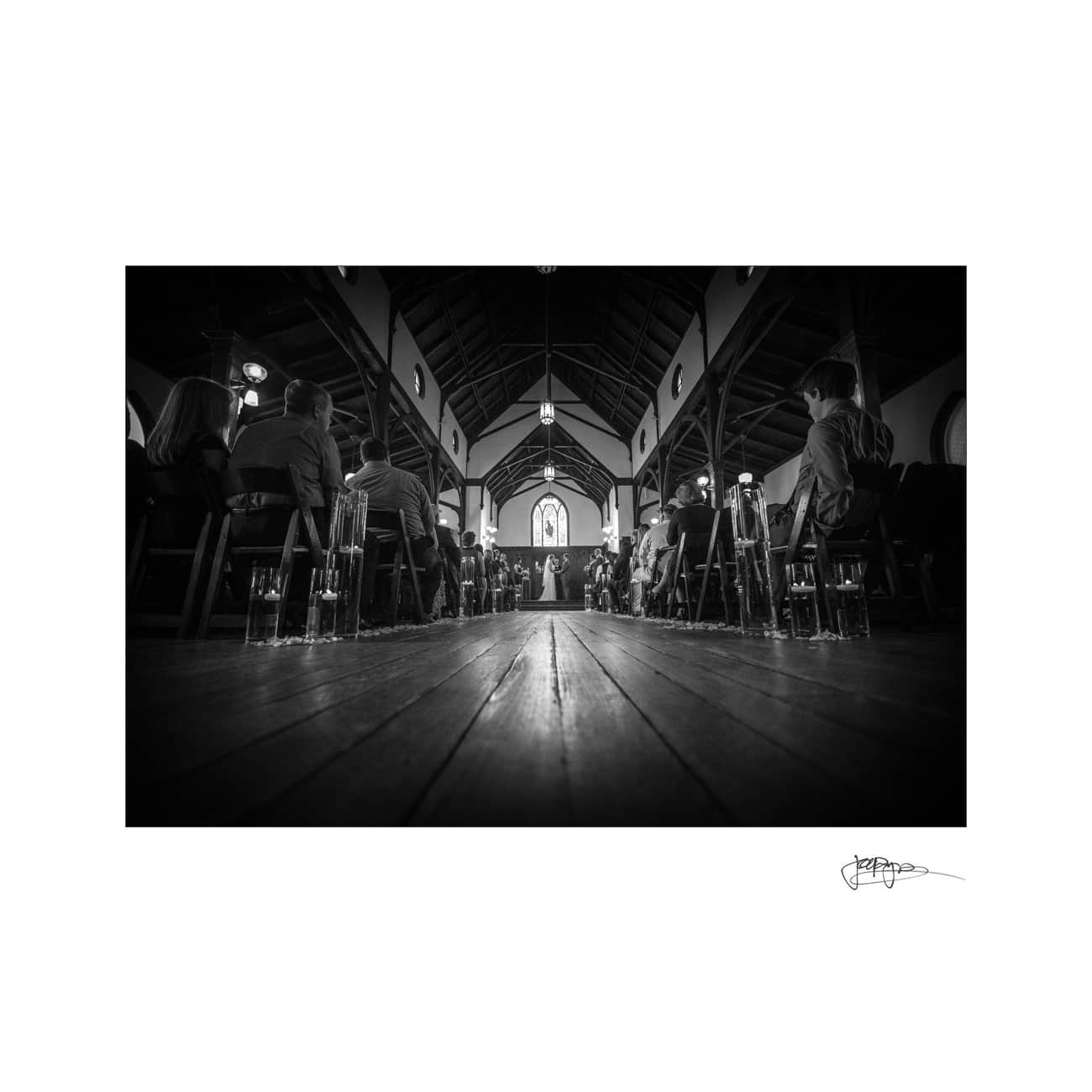 Black and white photo of a wedding ceremony in a church. The shot is taken from the floor, showing the bride and groom at the altar in the distance. Guests are seated along the aisle, with candles lining the path.