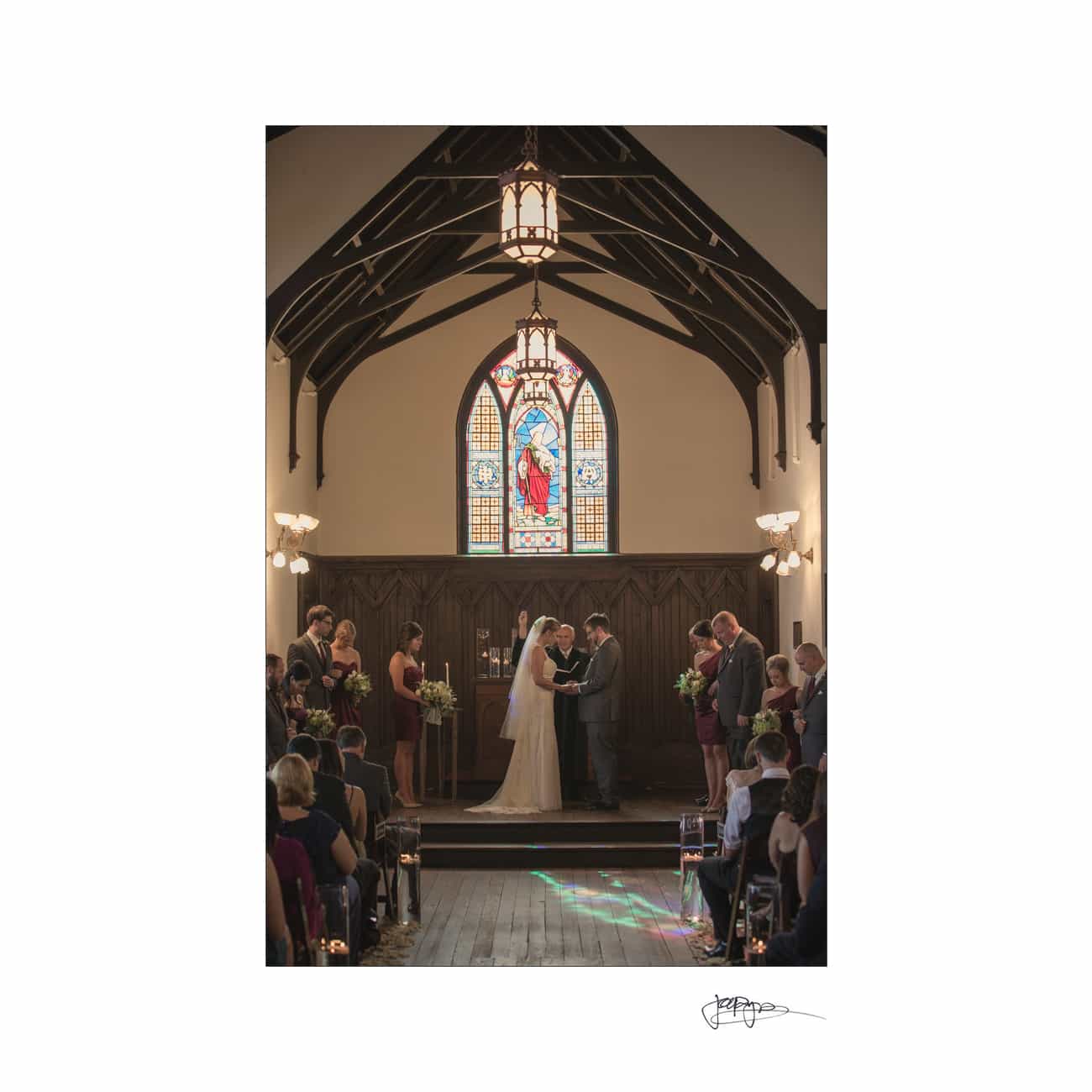 A couple stands at the altar during a wedding ceremony in a small chapel. The room features wooden beams and a colorful stained glass window above the altar. Guests are seated on either side.