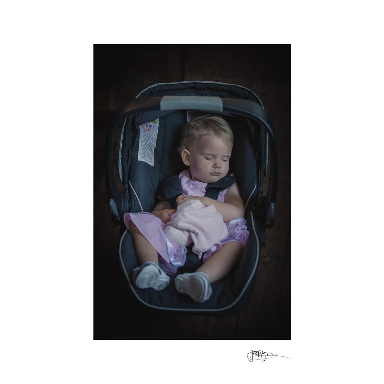 A toddler asleep in a car seat, wearing a pink dress and holding a soft toy. The car seat is dark, and the background is dimly lit, creating a peaceful atmosphere.