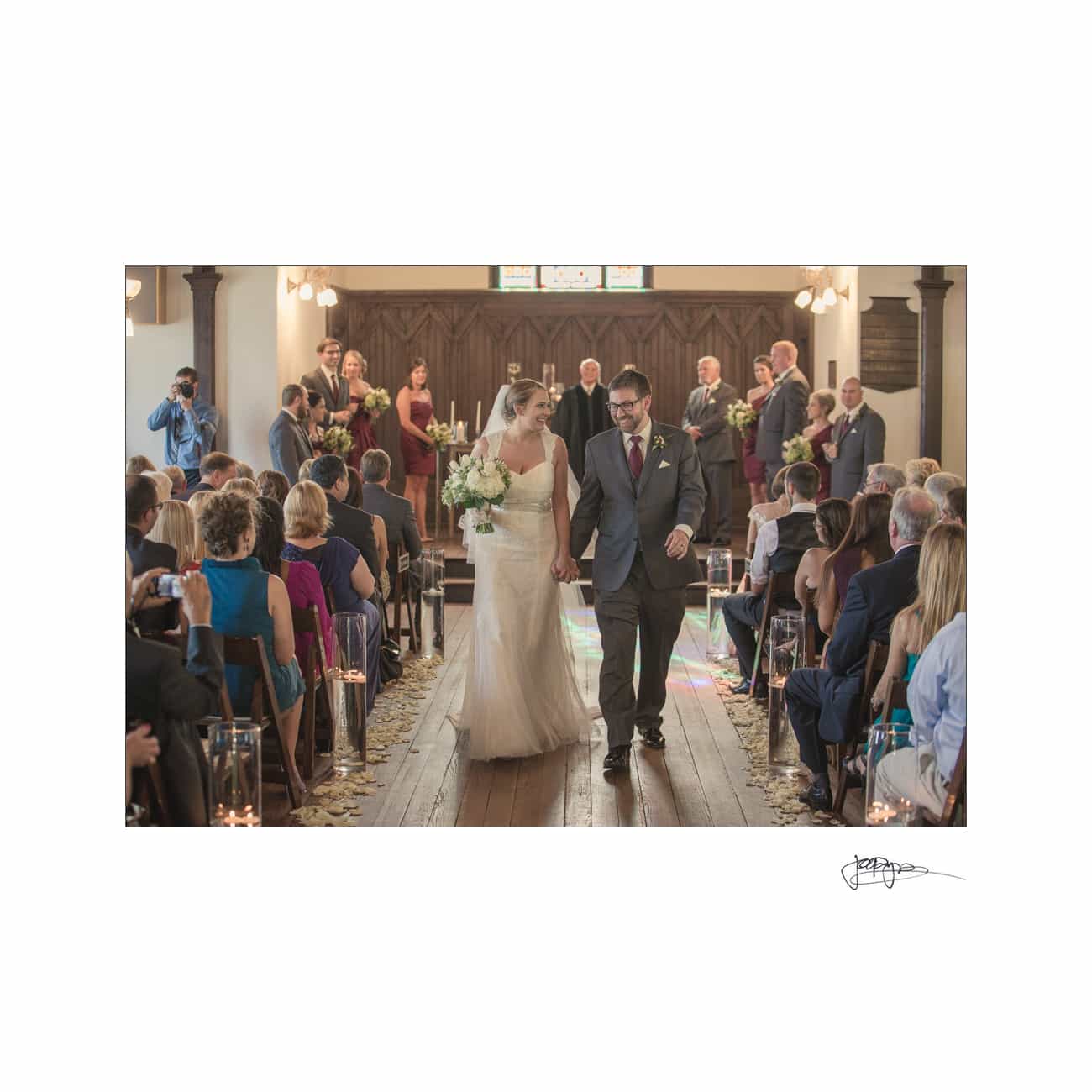 A bride and groom walk down the aisle in a rustic wedding venue. Guests seated on both sides look on, with flower petals scattered on the wooden floor. Photographers capture the moment.